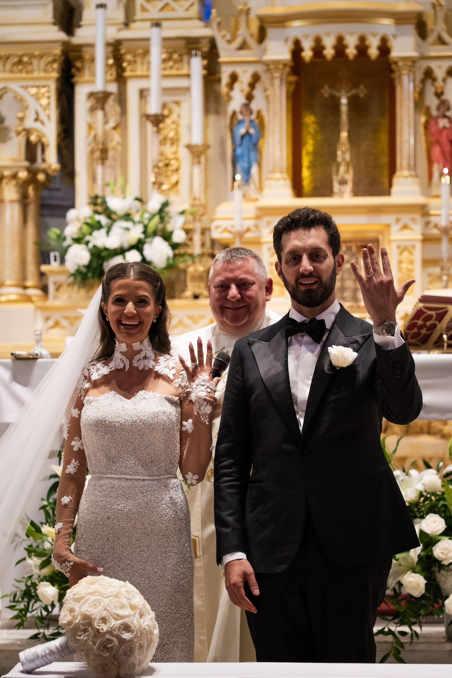 Newlyweds joyfully display wedding rings with smiling priest at ornate Catholic church altar.