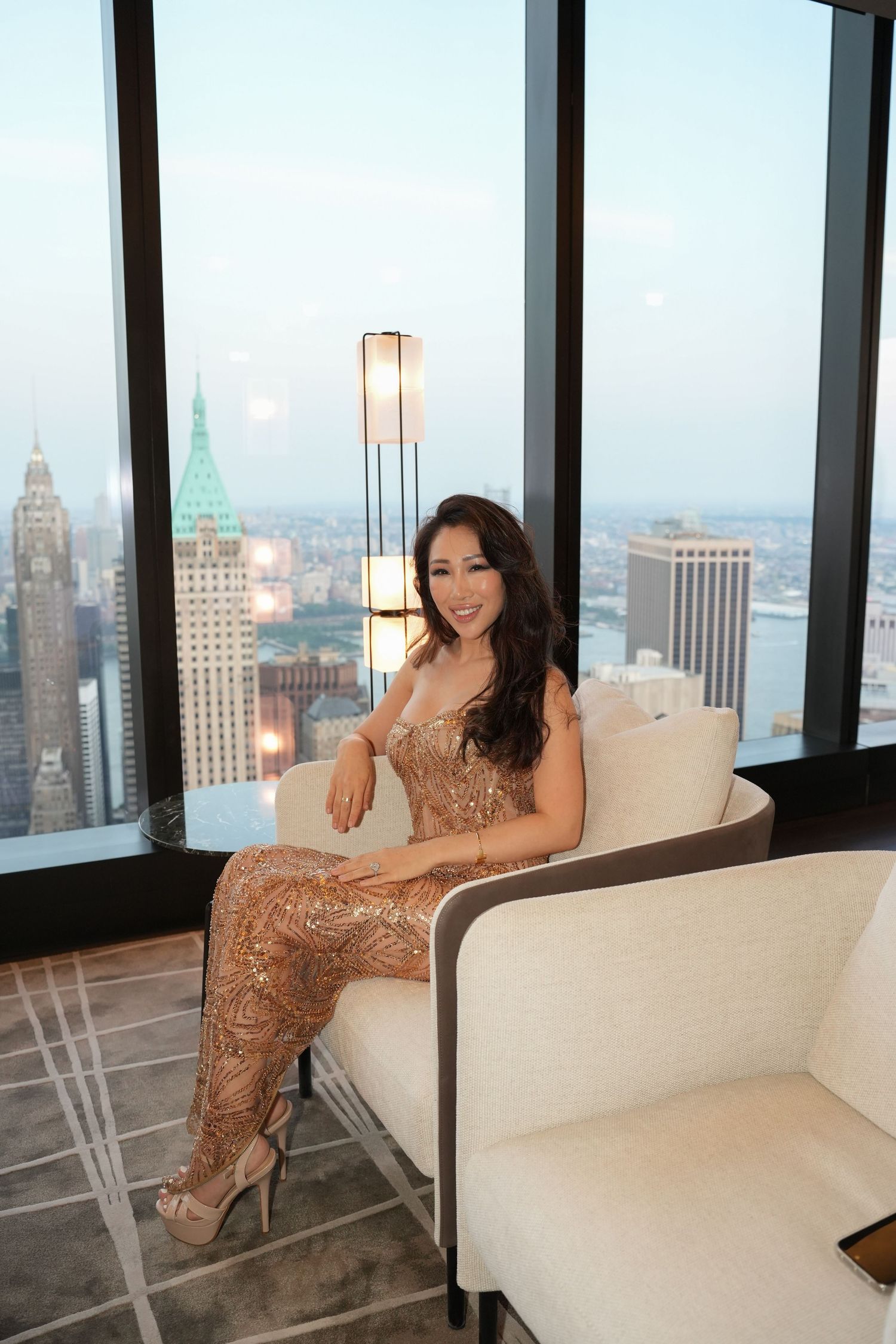 Woman in gold sequin gown posing on white sofa with panoramic New York City skyline view.