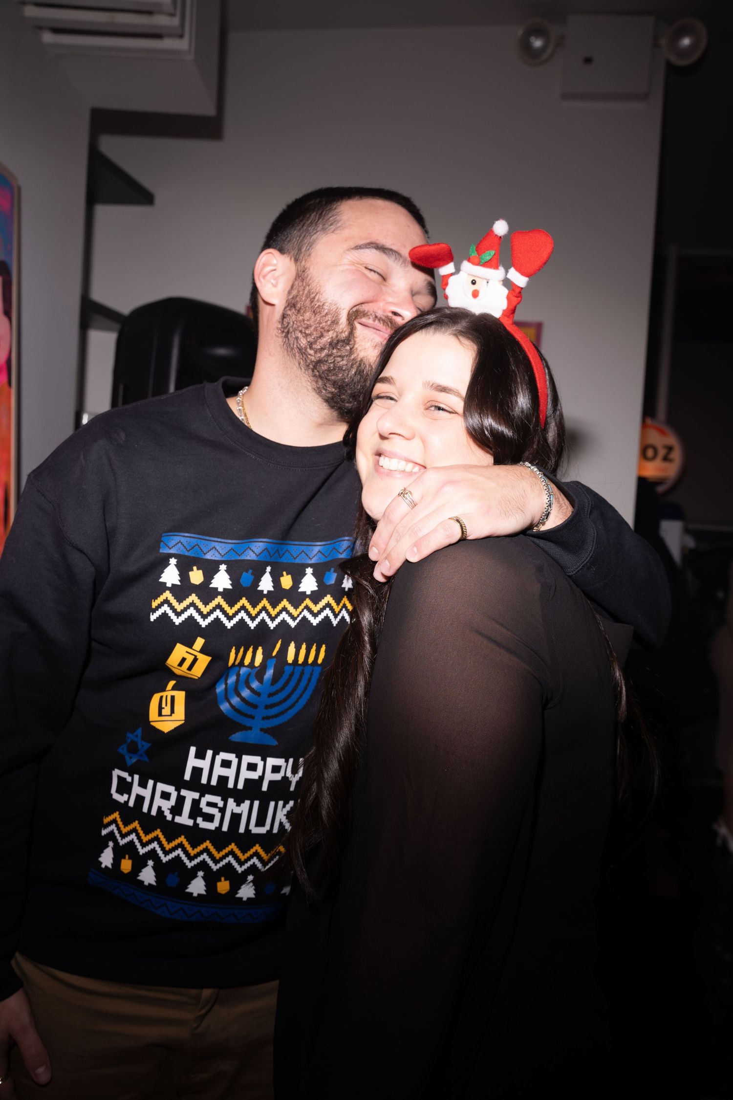 Couple embracing and laughing at a holiday party, man wearing Happy Chrismukkah sweater.