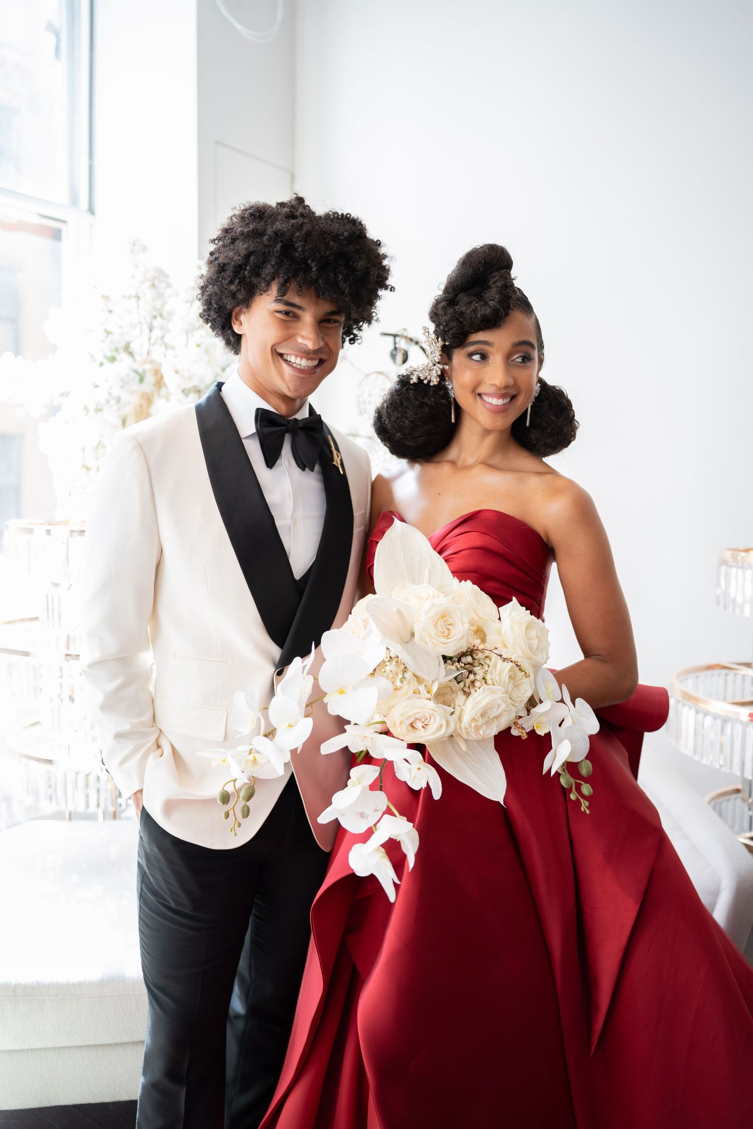 Joyful wedding couple standing together, groom in white tuxedo, bride in red ball gown holding white floral arrangement indoors.