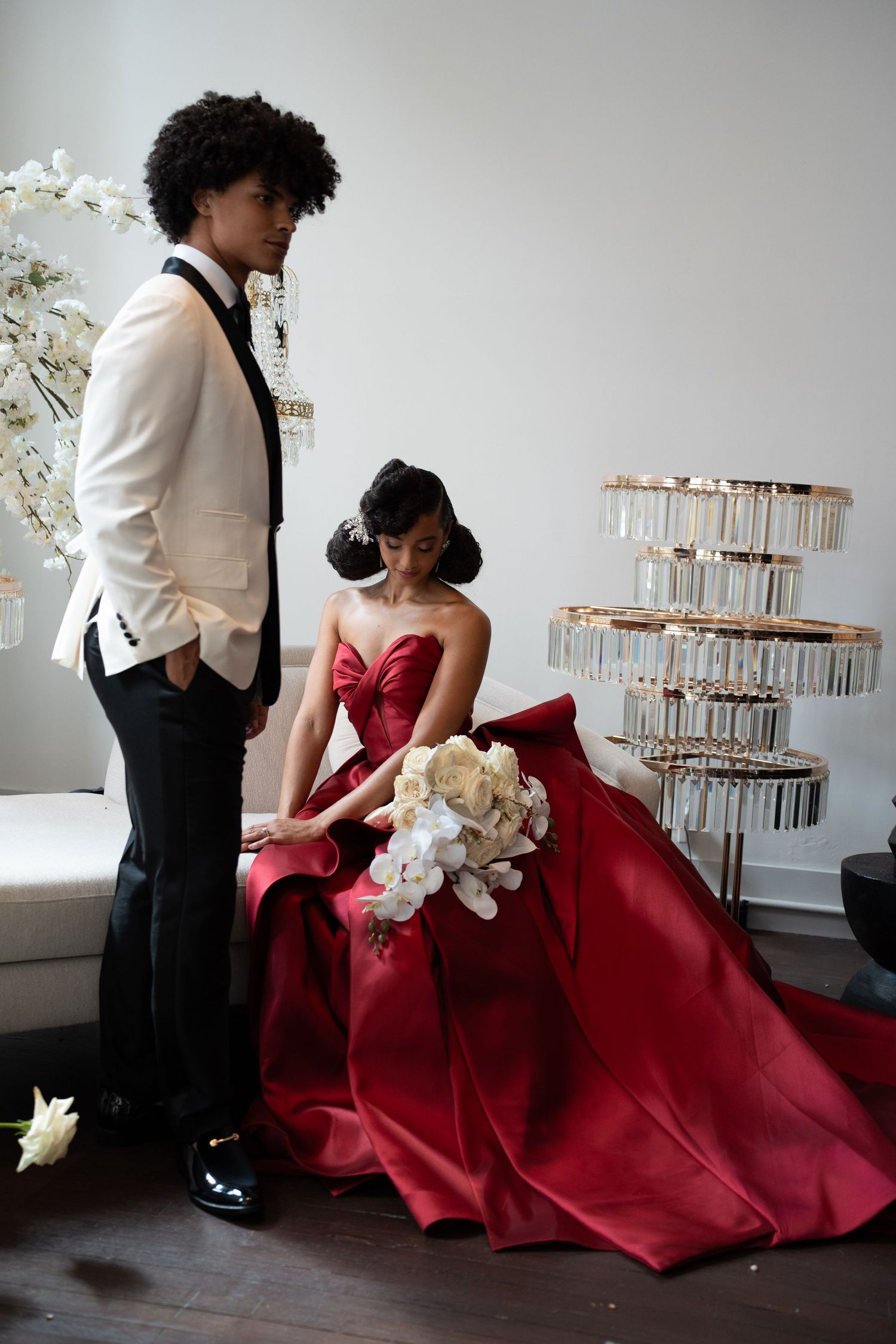 Bride in dramatic red ball gown holding white orchid bouquet seated while groom in white tuxedo stands nearby.