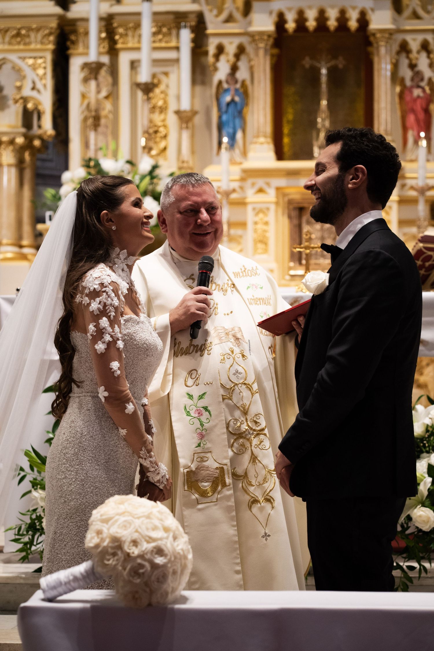 Bride and groom exchange vows with smiling priest officiant at ornate Catholic church altar during wedding ceremony.