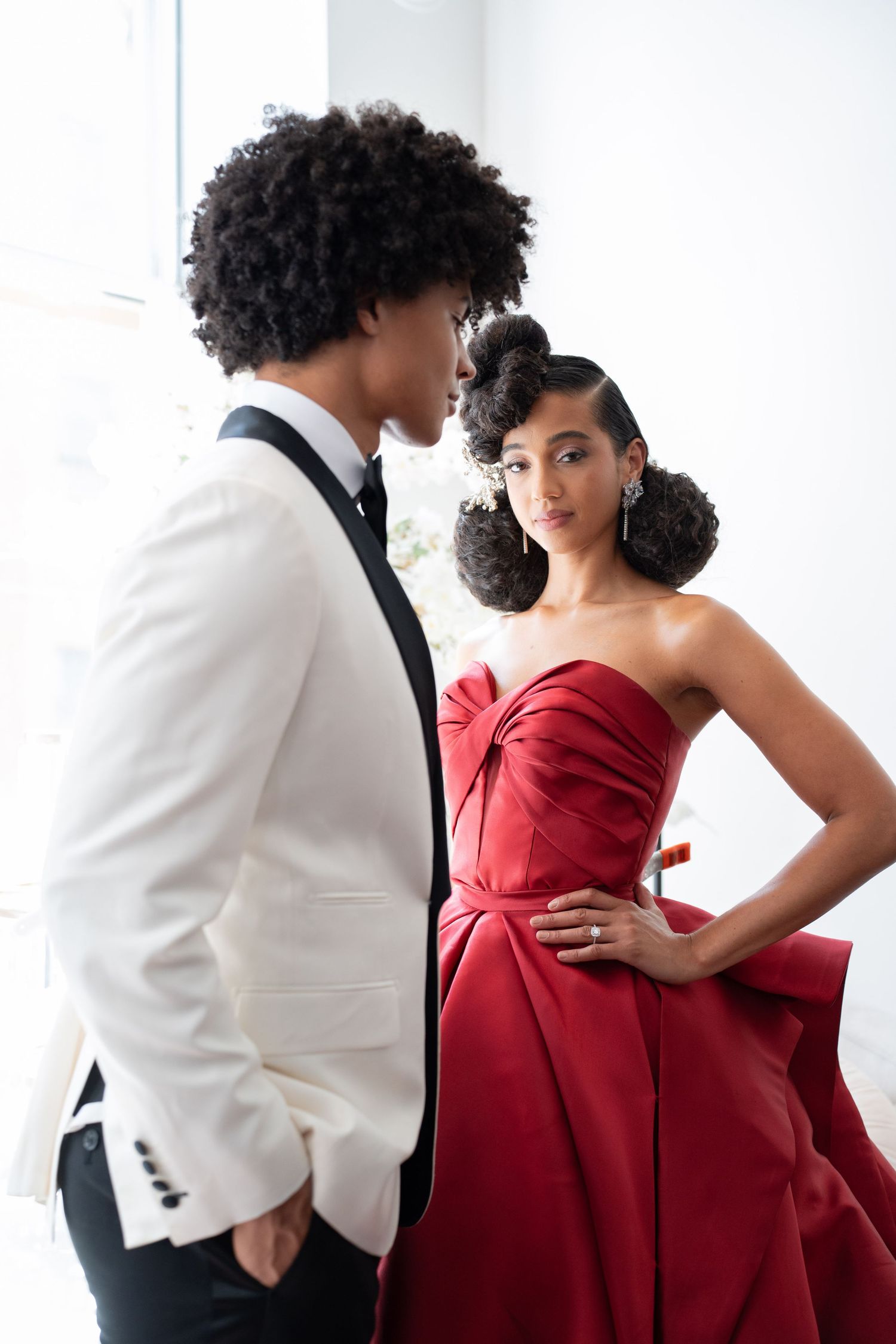 Beautiful bride in red strapless ballgown with hand on hip facing groom in white tux.