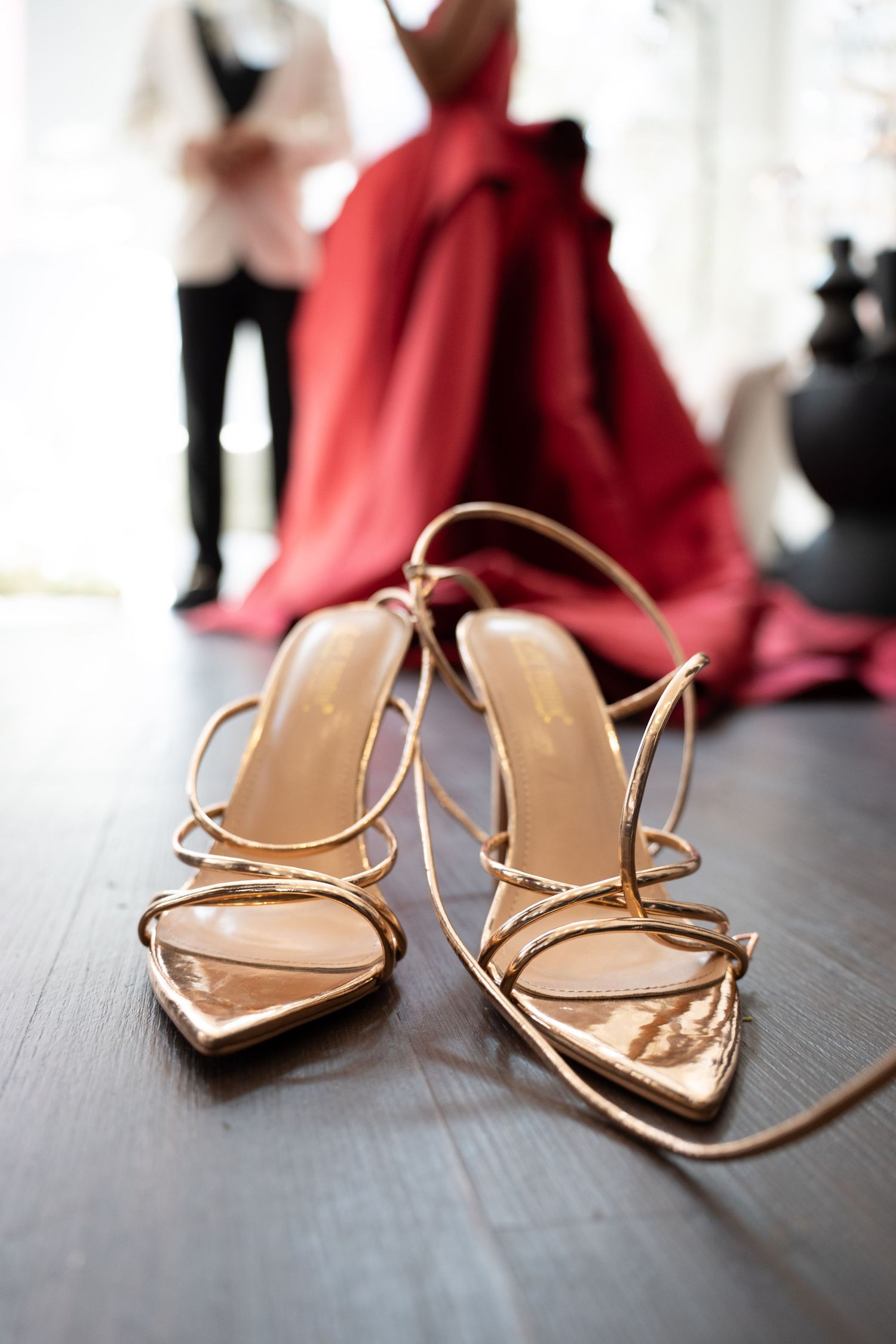 Gold strappy heeled sandals on wooden floor with red bridal gown in background.