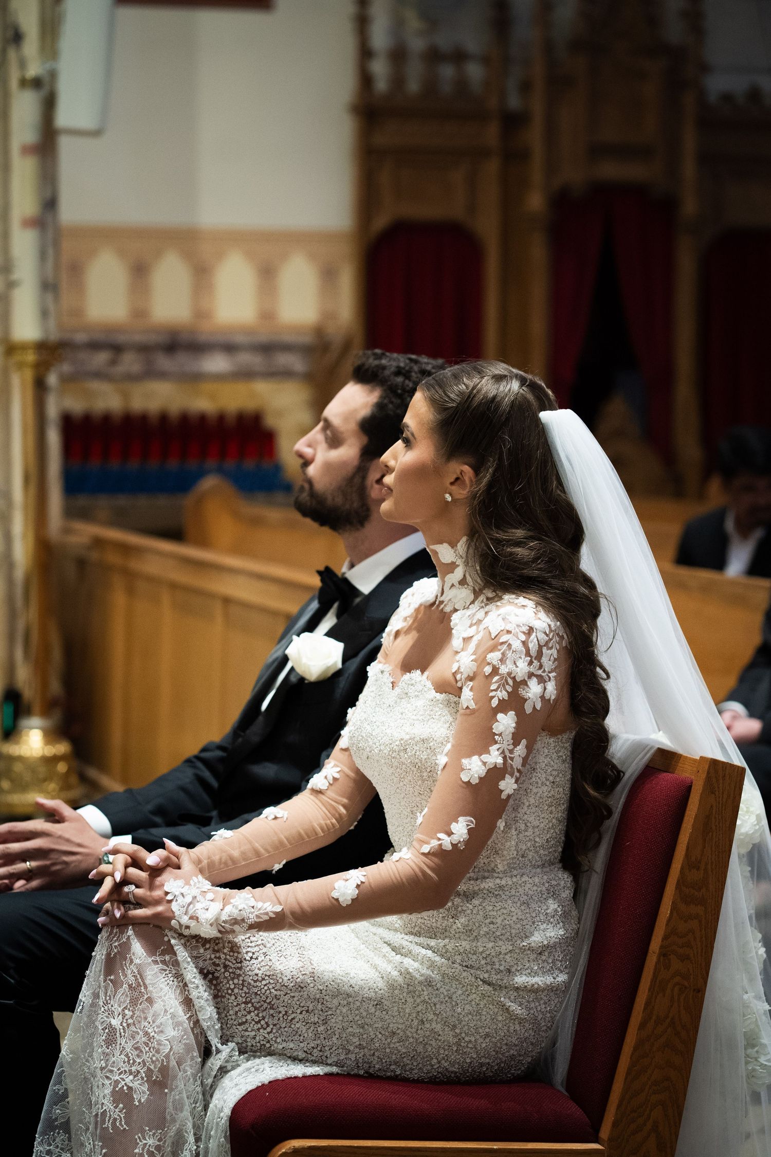 Bride in floral lace gown and groom in tuxedo sit together during church wedding ceremony.