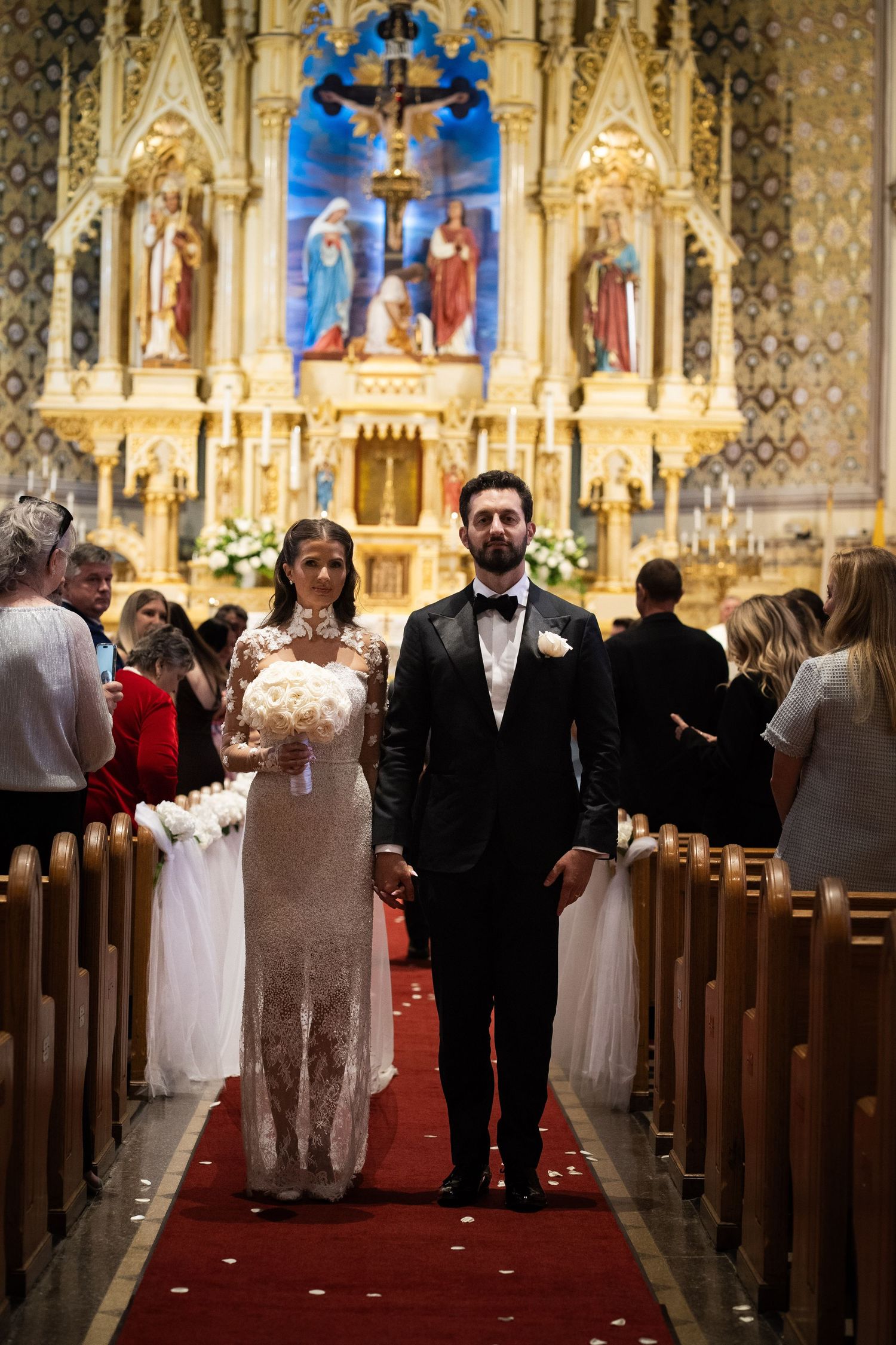 Newlyweds walk down red carpet aisle of ornate Catholic church after wedding ceremony.