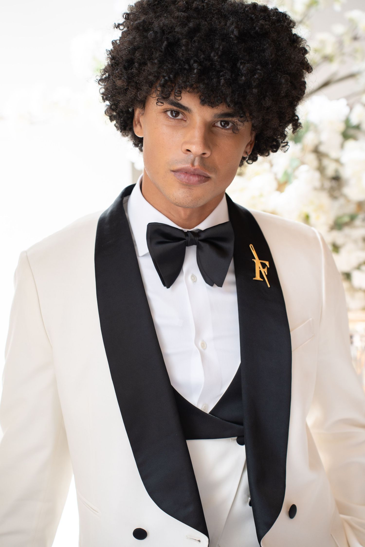 Close-up portrait of groom in white tuxedo with black bow tie, vest, and gold decorative lapel pin.