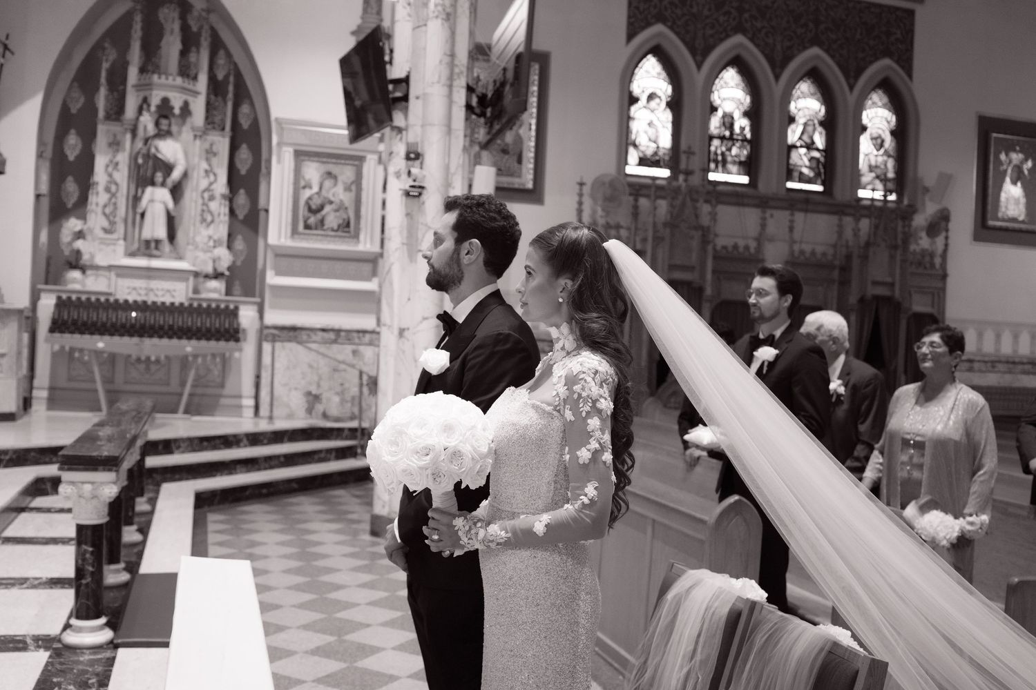 Bride in lace gown with long veil and groom in tuxedo stand together at the church altar during their wedding ceremony.