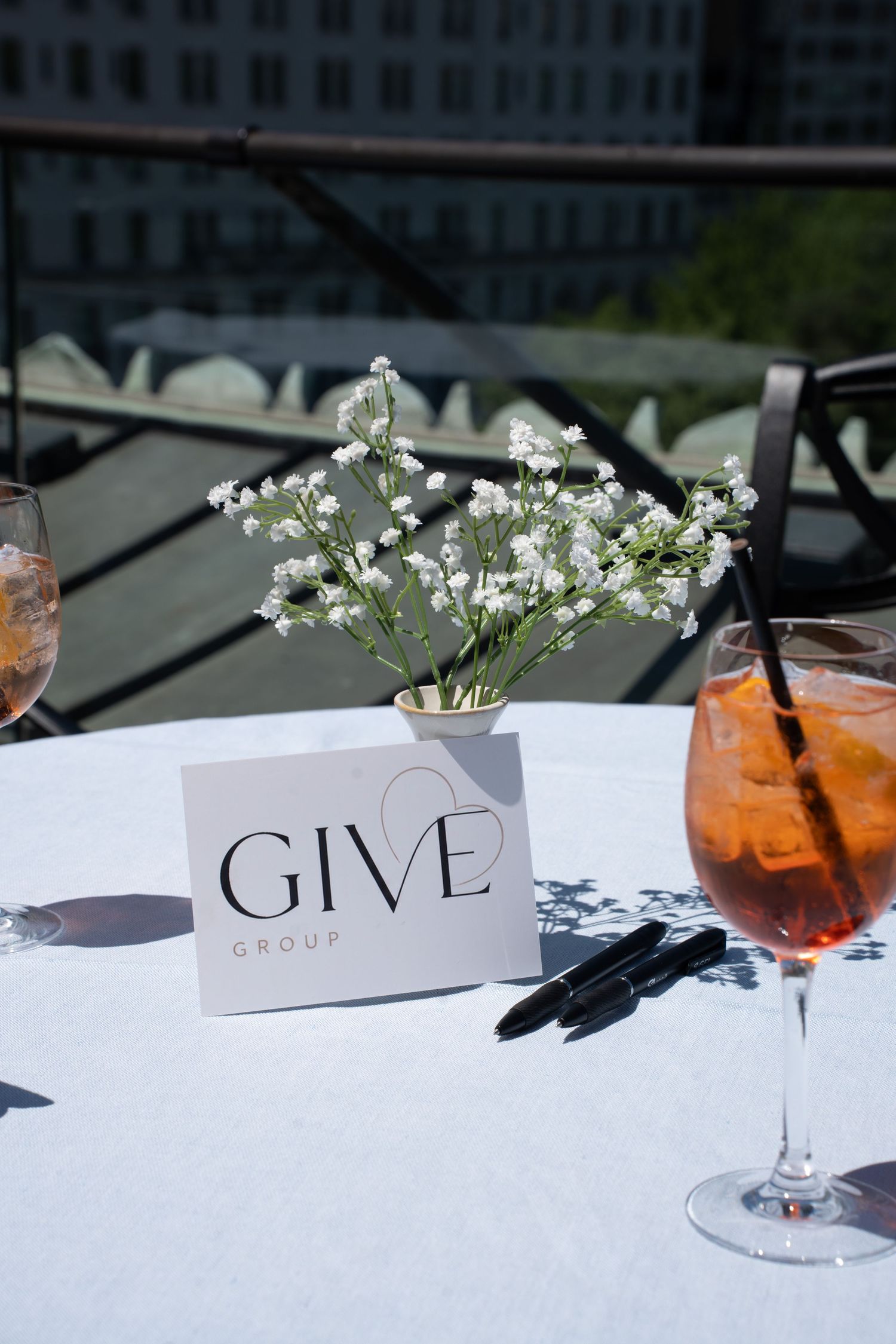 GIVE Group branded card displayed on white tablecloth with baby's breath flowers and Aperol Spritz.