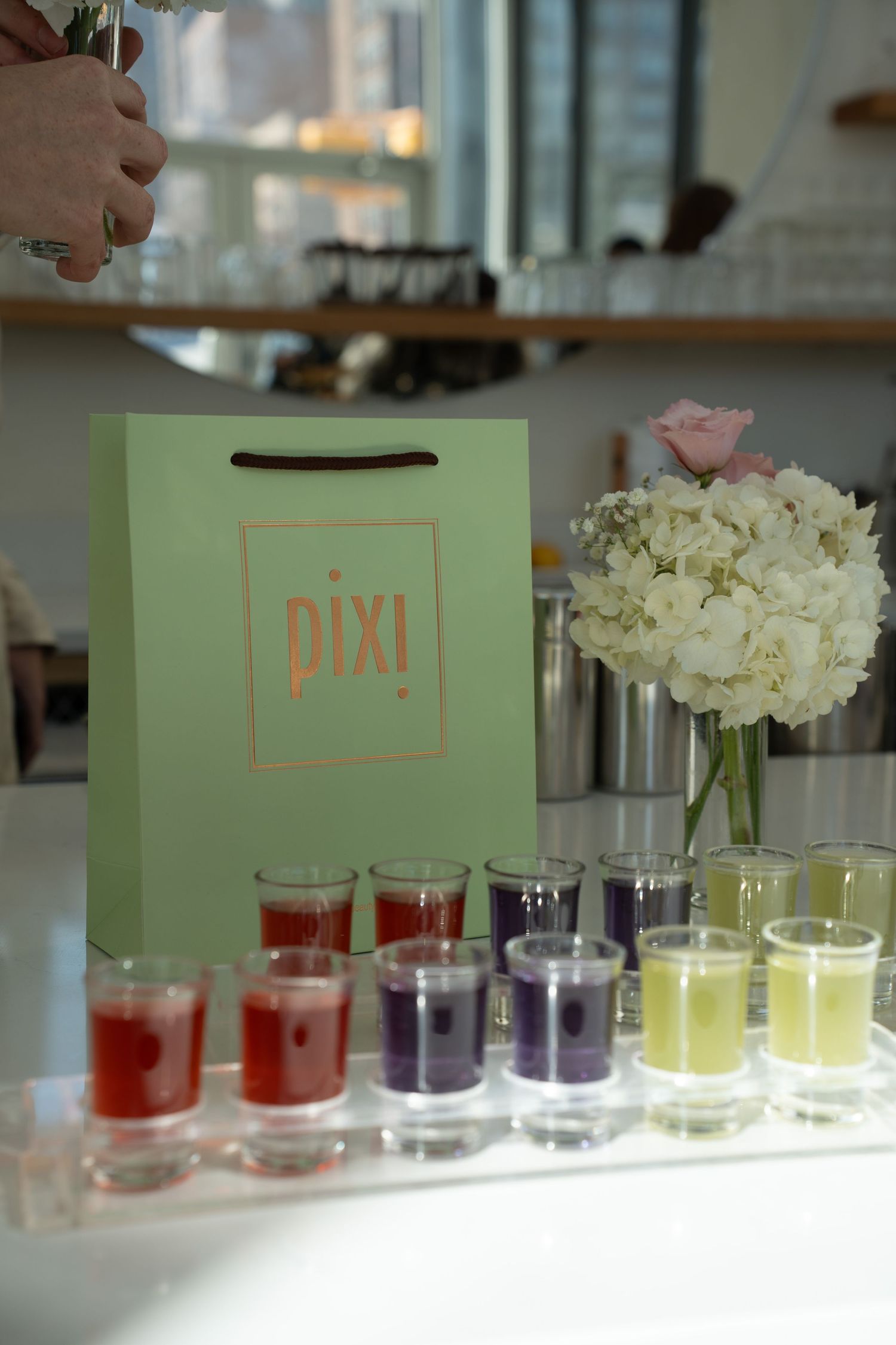 Mint green Pixi gift bag displayed beside colorful shot glasses and white hydrangea flowers.