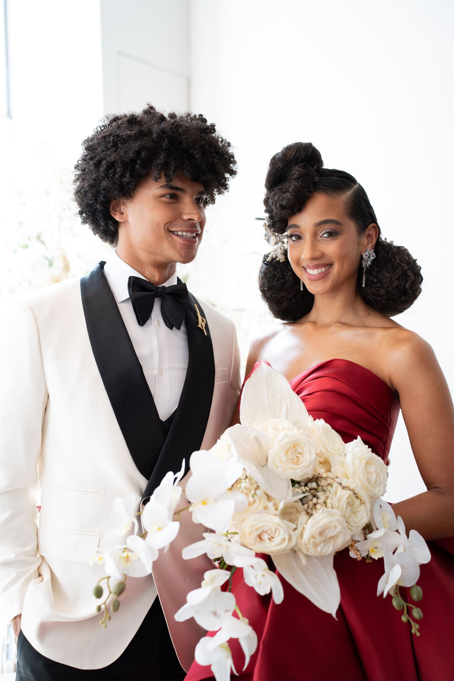 Smiling wedding couple, groom in white tuxedo with bow tie, bride in red strapless gown holding white roses and orchids bouquet.