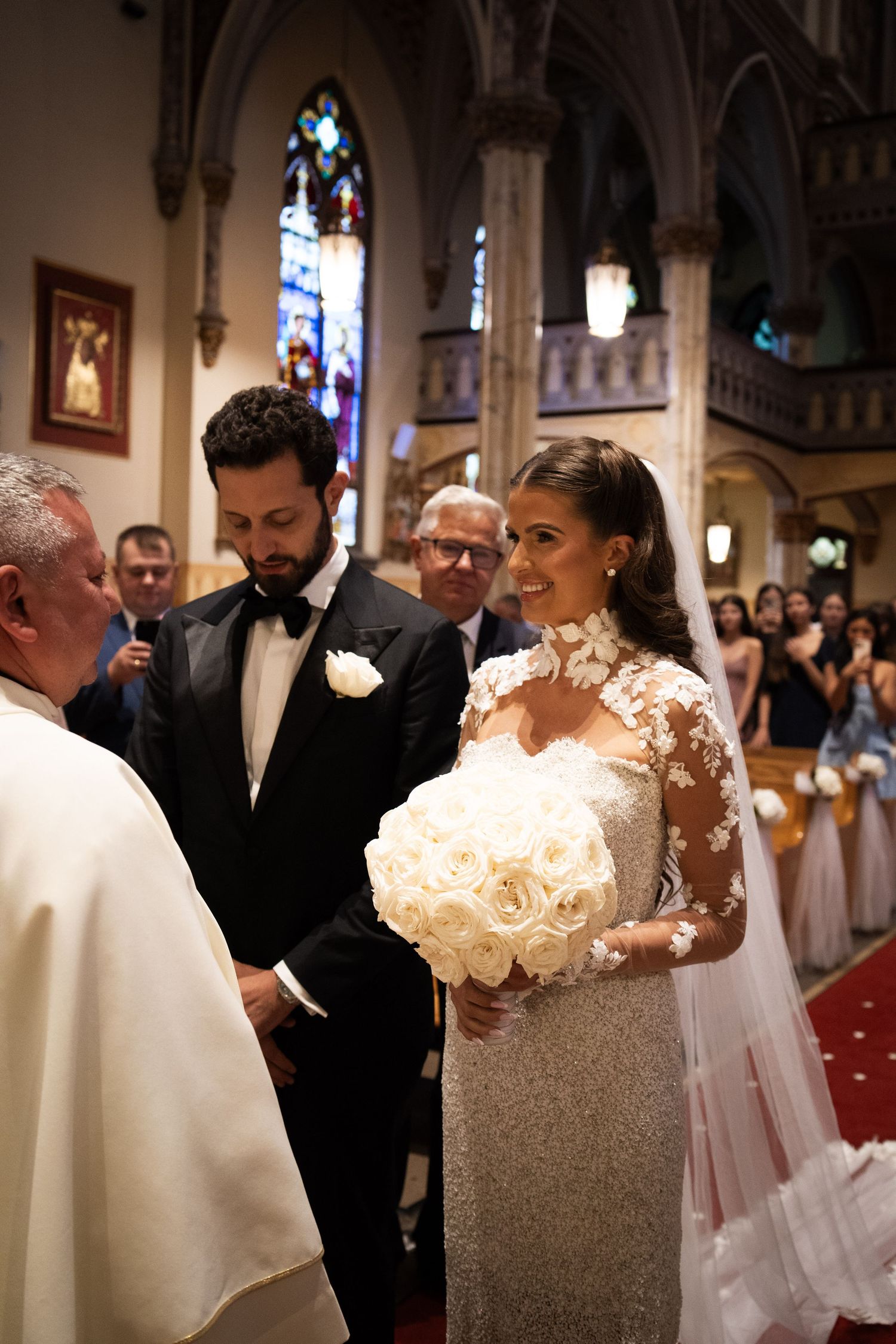 Bride in lace wedding gown holding white rose bouquet stands with bearded groom before priest at ceremony.