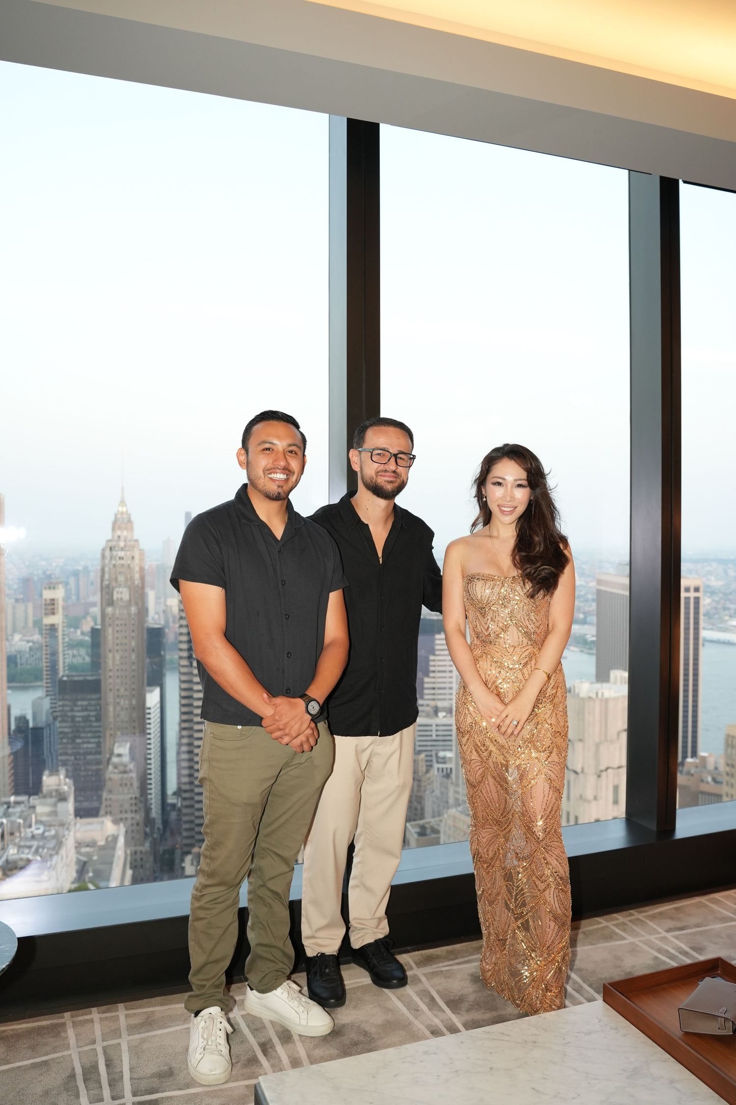 Three elegantly dressed people posing together in a high-rise luxury apartment with city skyline views.