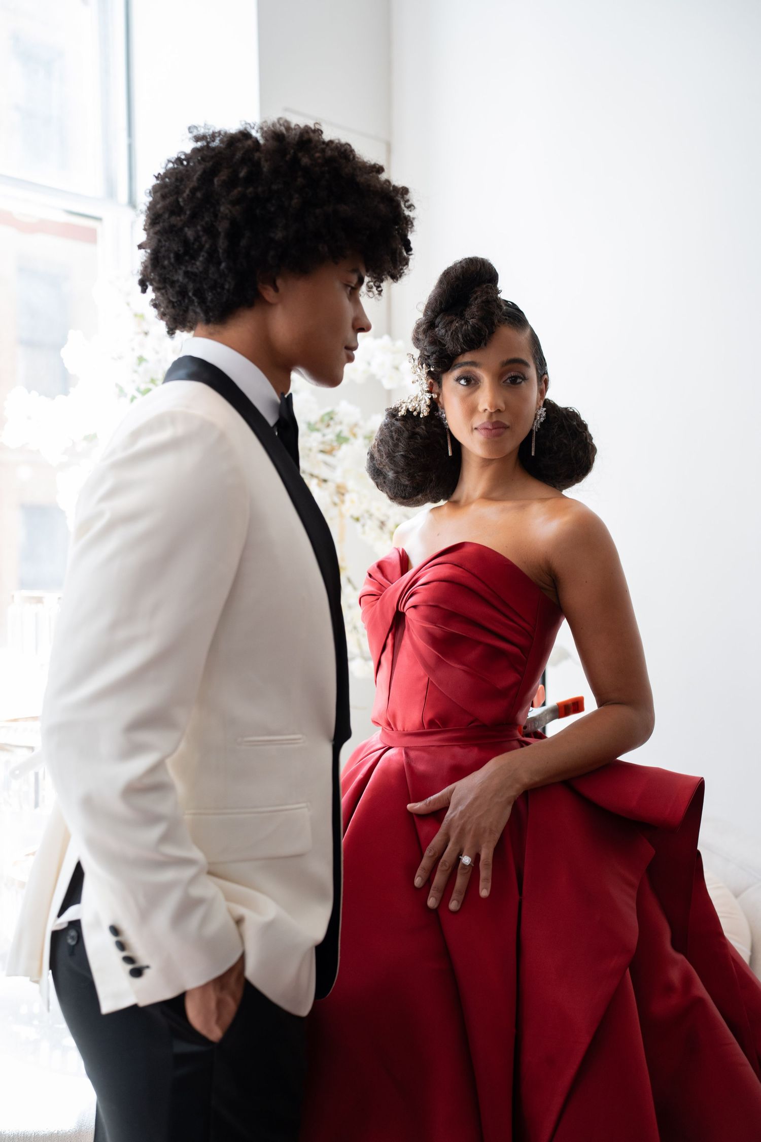 Stylish bride in red strapless gown gazing at groom in white tuxedo jacket.