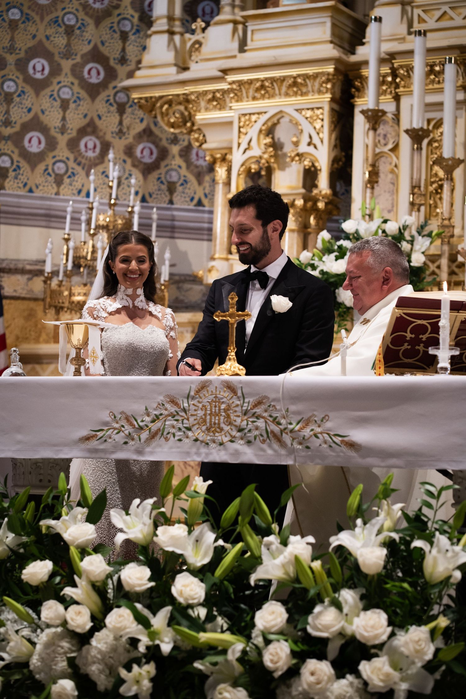 Bride and groom sign wedding documents at flower-decorated altar with priest in Catholic church.