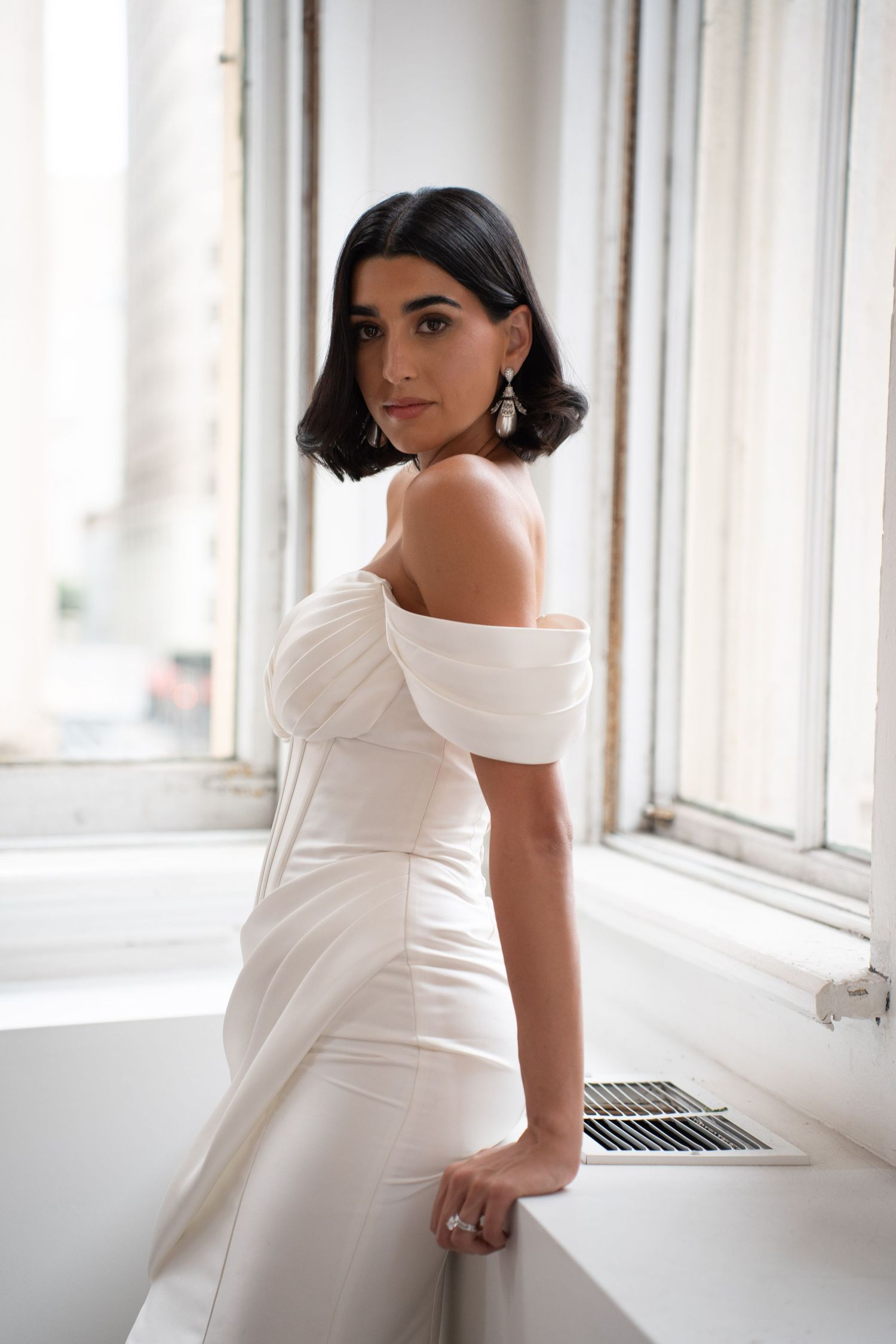 Bride in off-shoulder white gown poses elegantly by window, showcasing draped bodice and drop earrings.