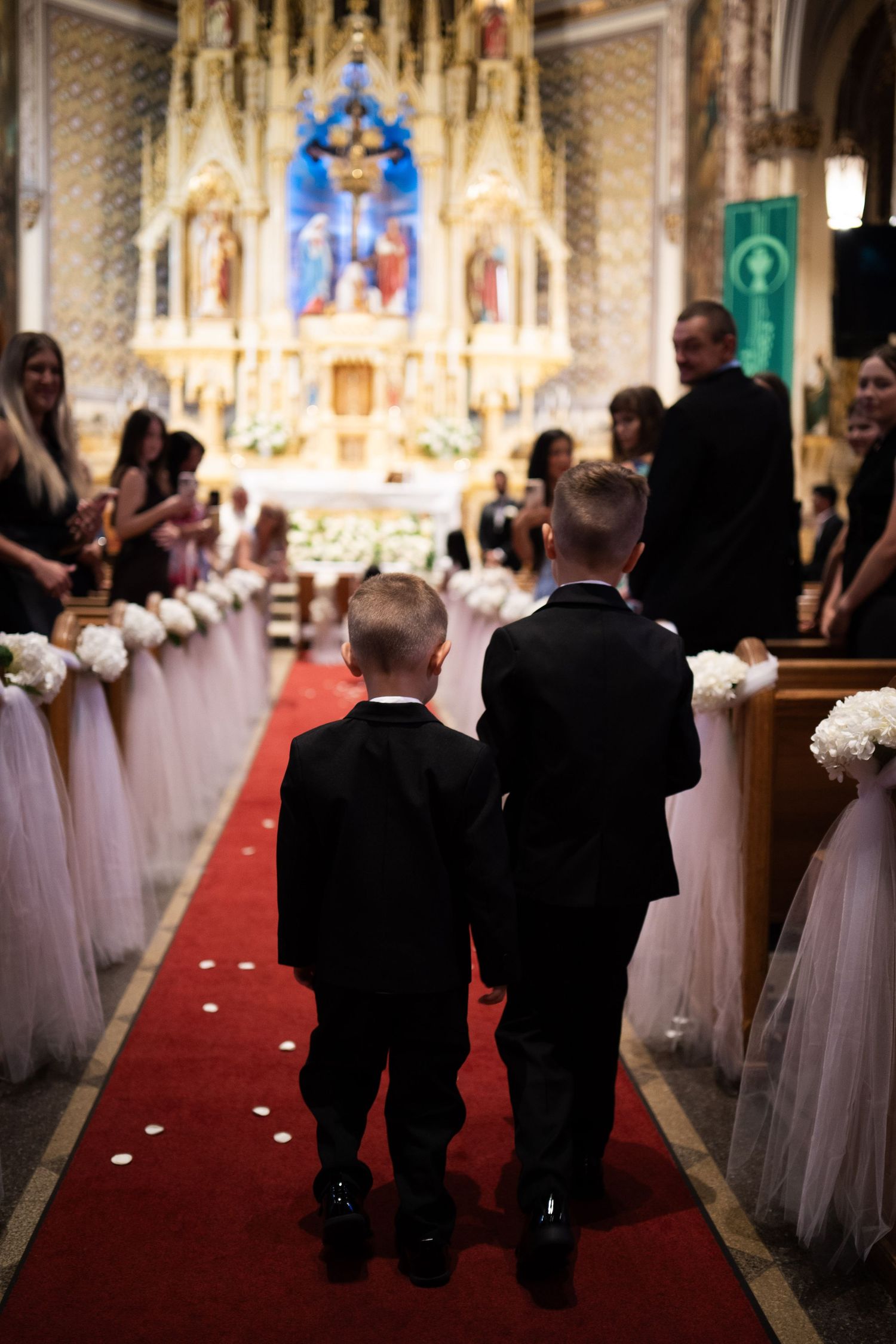Two young ring bearers in black suits walk down a red carpet aisle in an ornate Catholic church during a wedding ceremony.