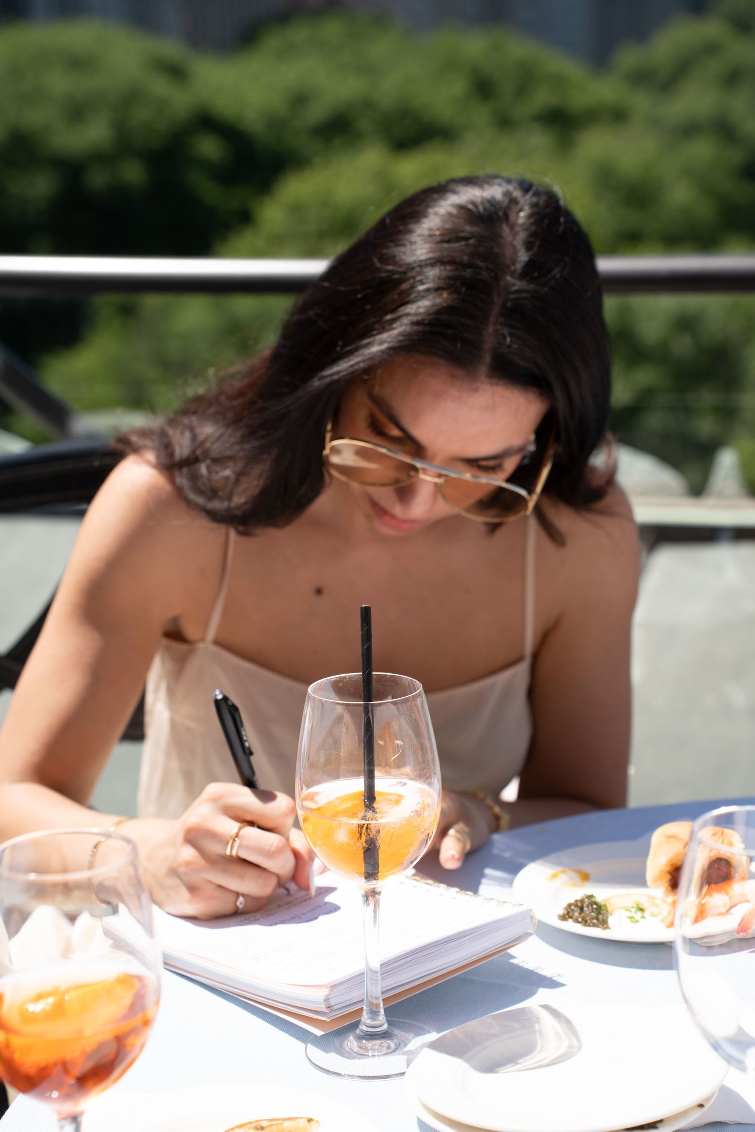 Dark-haired woman in sunglasses writing in notebook beside Aperol Spritz at outdoor NYC brunch.