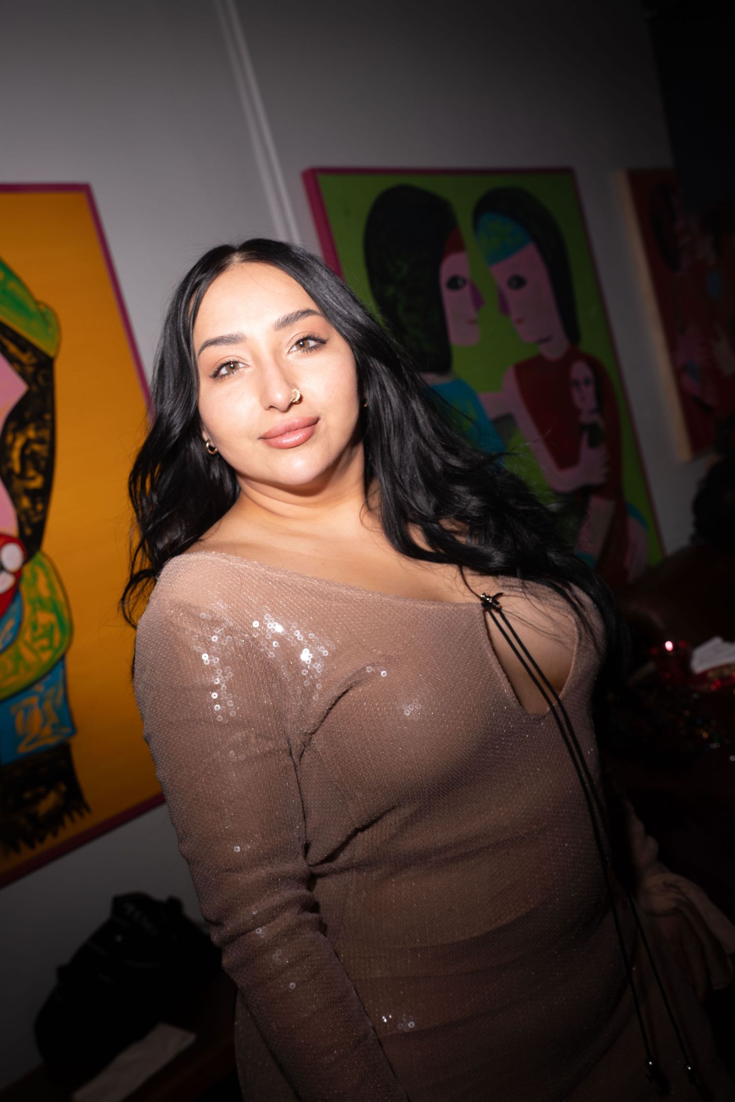 Young woman in sparkly beige top poses in front of colorful art gallery.