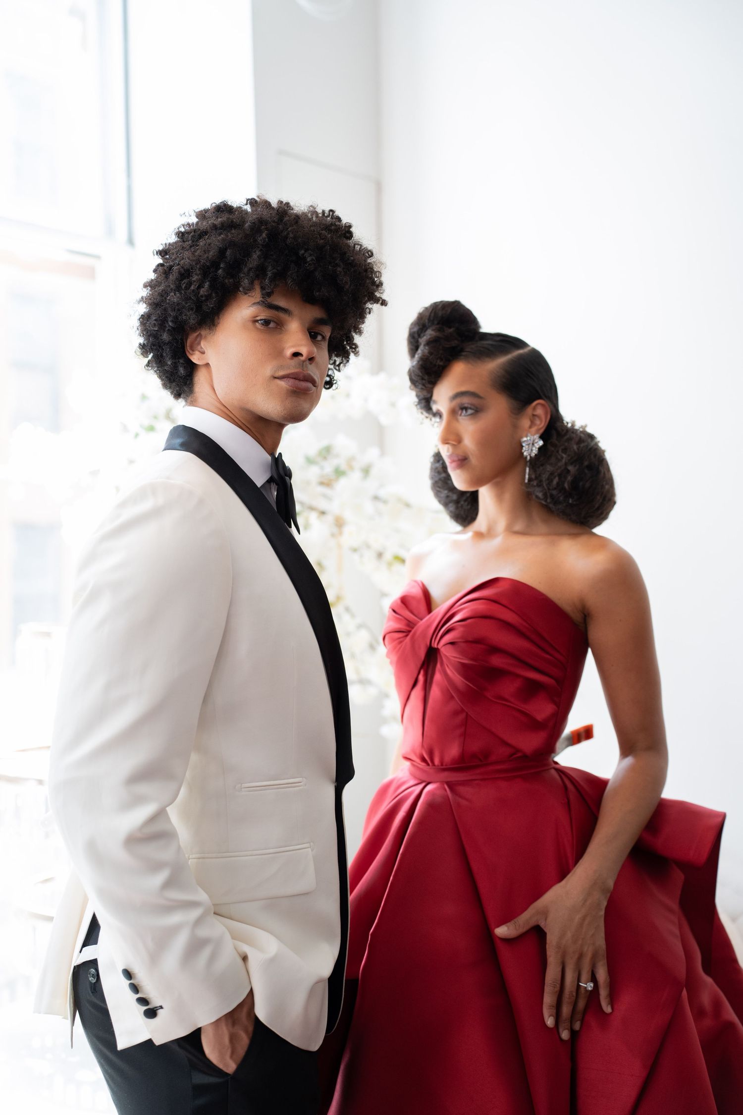 Elegant wedding couple in white tuxedo and red strapless gown standing by window.