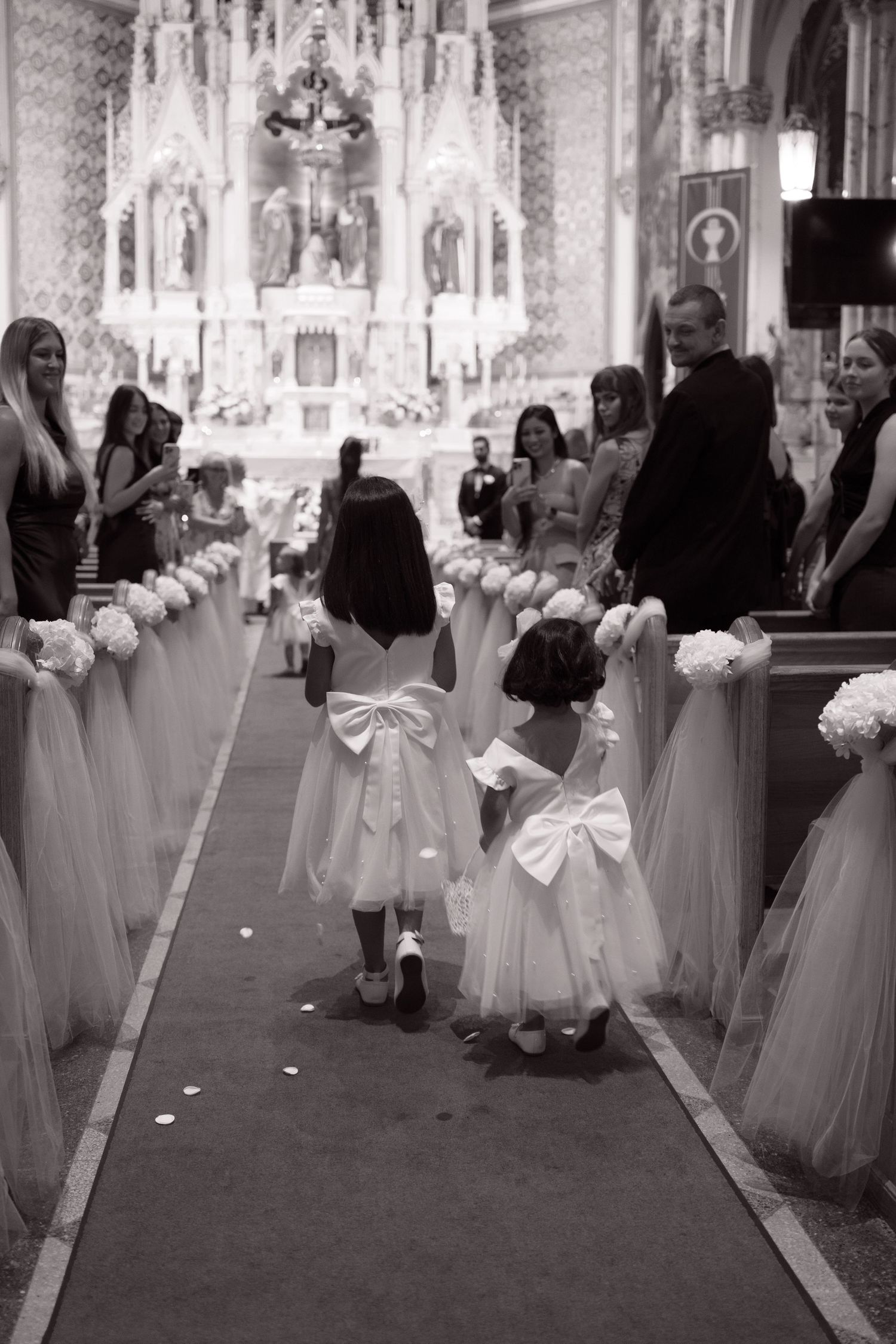 Two flower girls in white dresses with bows walk down a decorated church aisle during a wedding ceremony.