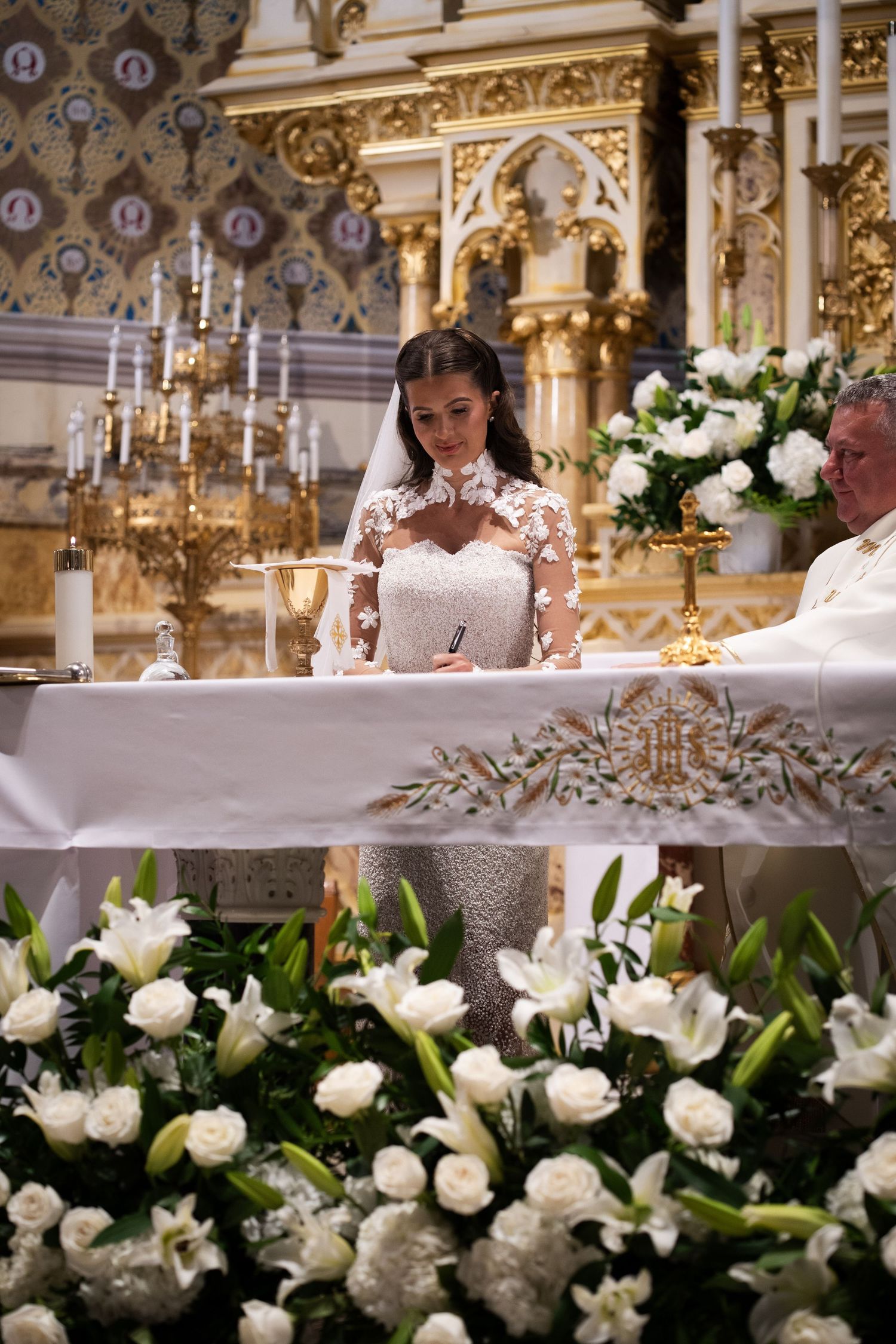 Bride in lace gown signs wedding register at flower-adorned altar in Catholic church.