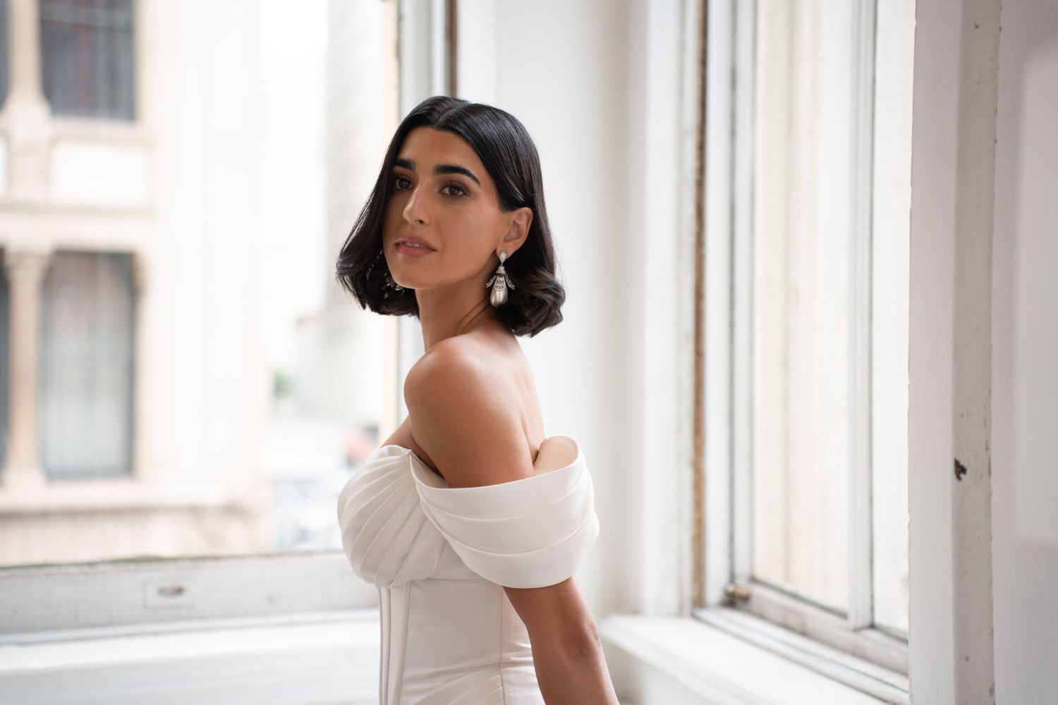Bride in draped off-shoulder white gown looks back over shoulder near bright windows wearing statement earrings.