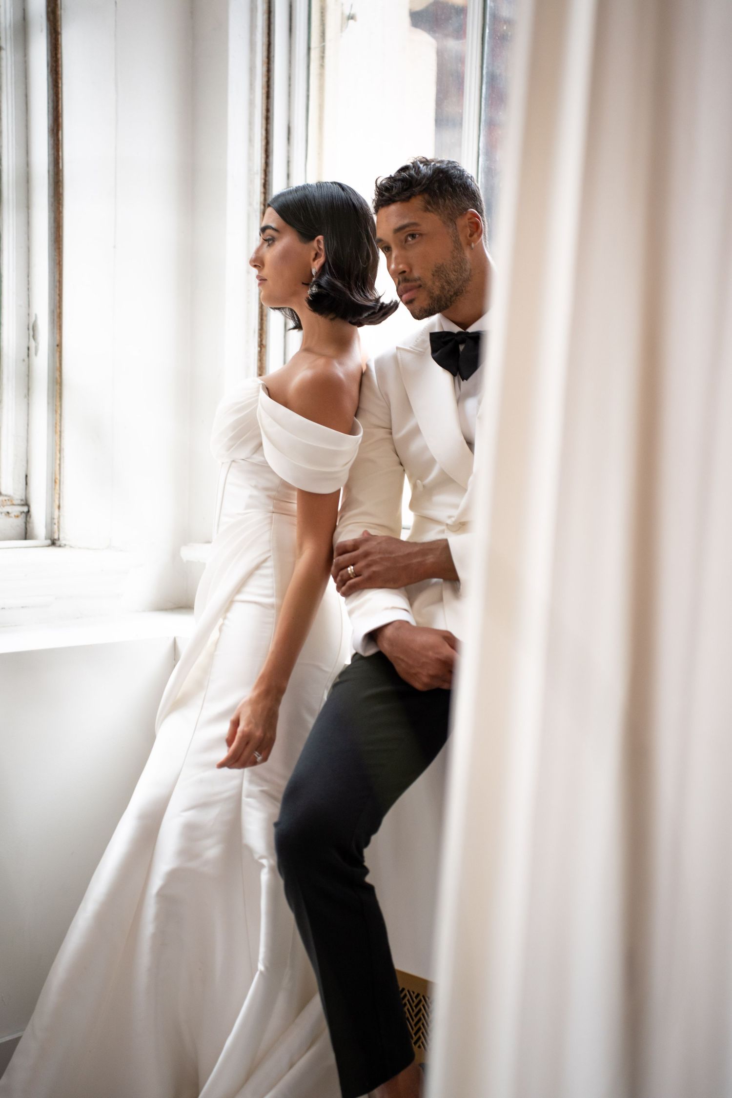 Bride and groom in wedding attire sharing intimate moment by window, groom in white tuxedo.