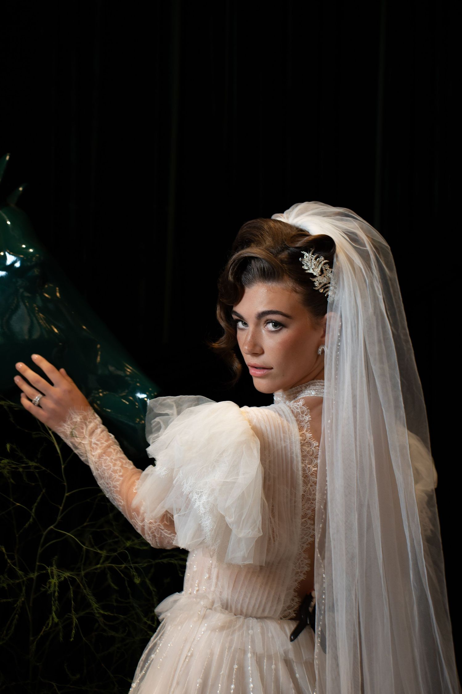 Elegant bride in lace wedding dress with long veil and hair accessory poses near dark foliage.