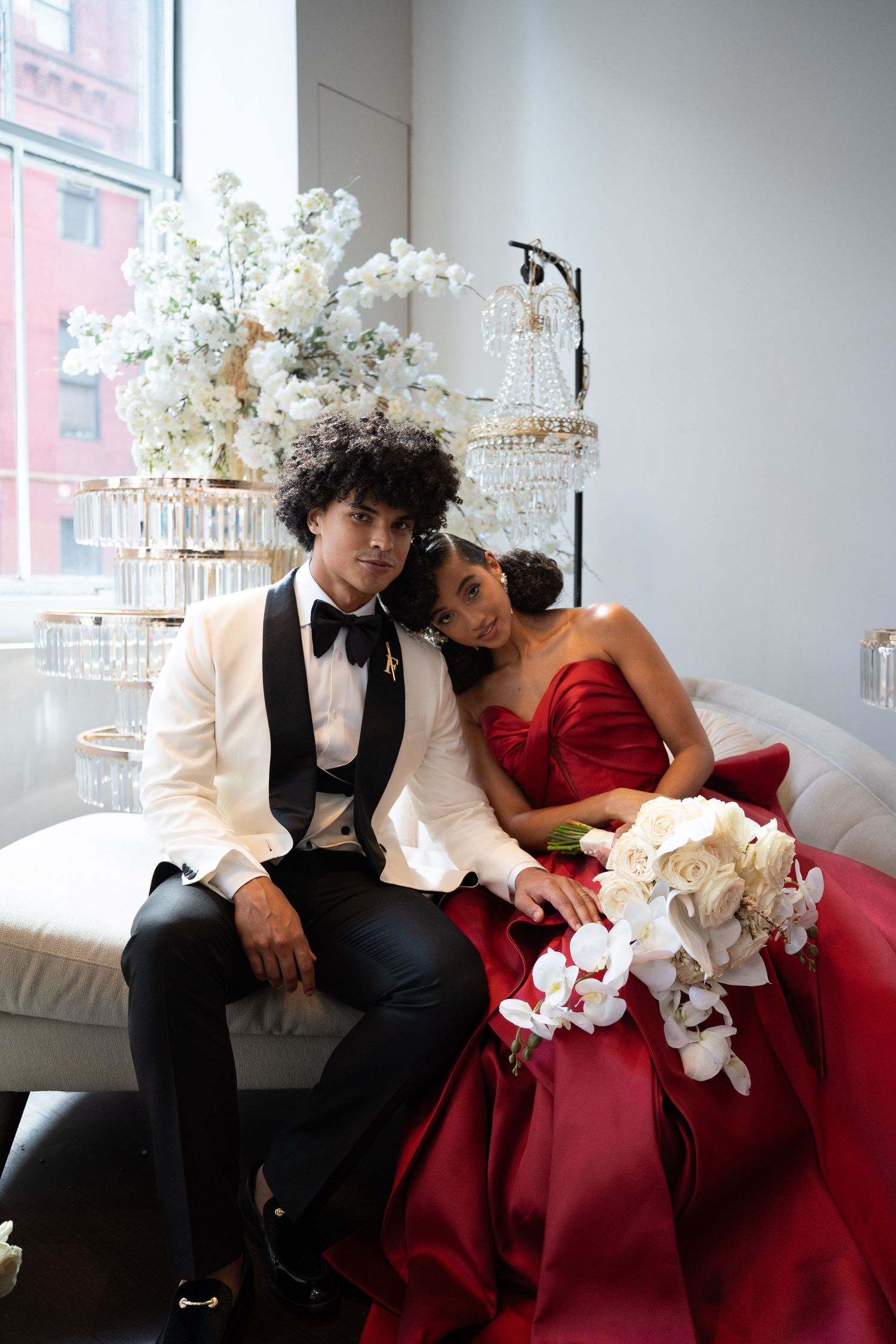 Bride in red gown and groom in white tuxedo pose together on sofa at elegant floral wedding venue.