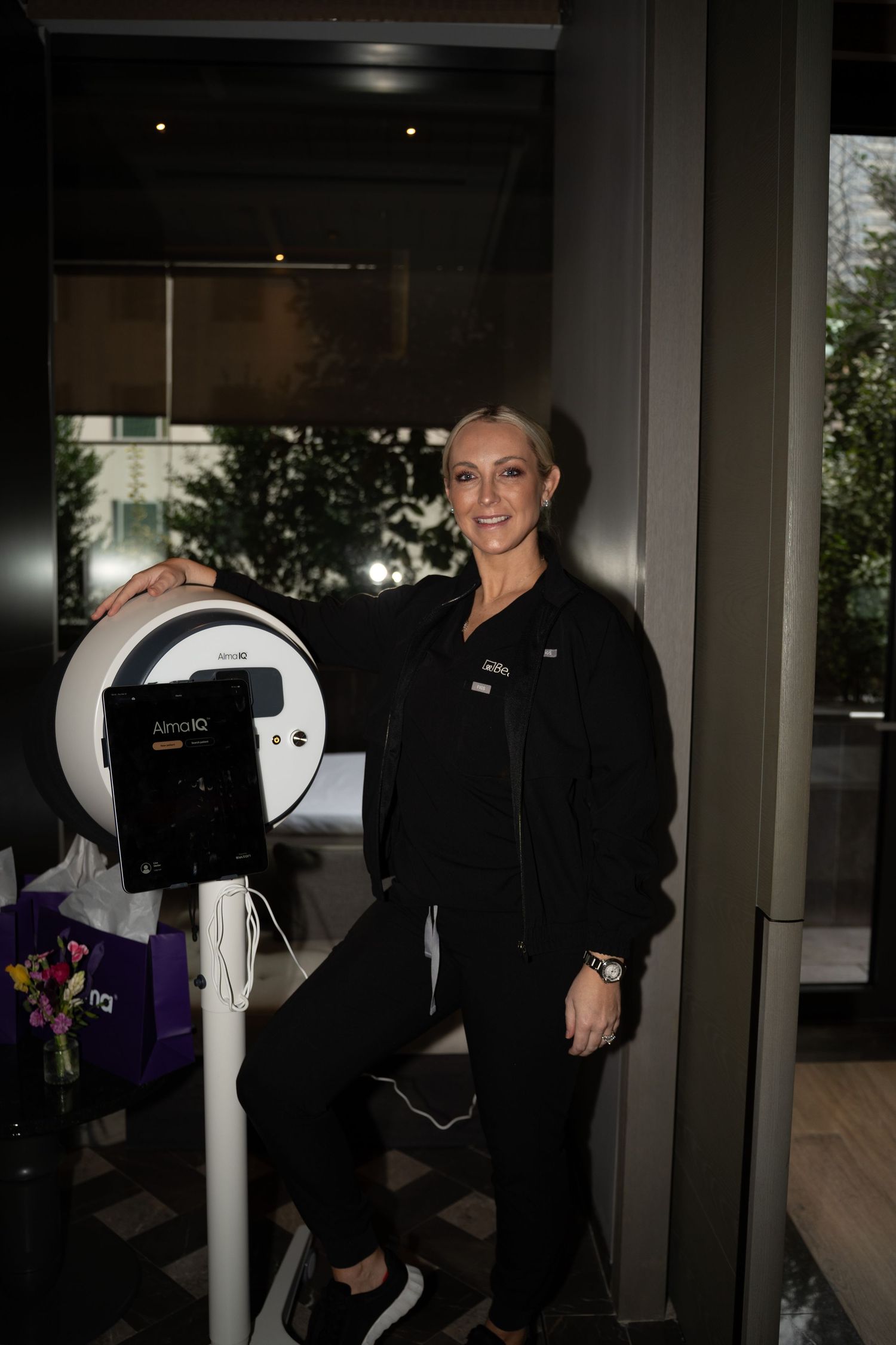 Smiling blonde woman in black uniform standing beside an Alma IQ skin analysis device at a spa event.