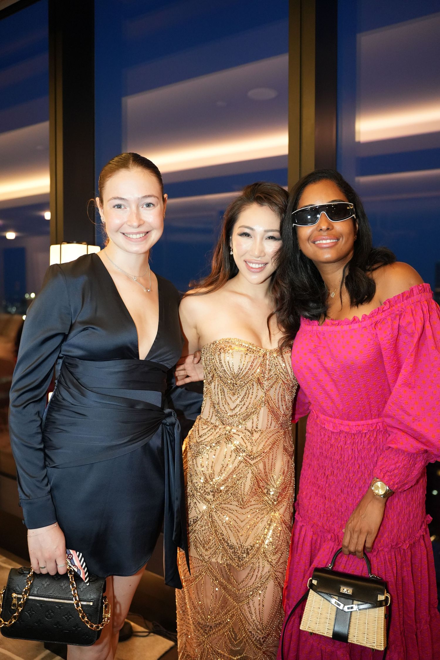 Three elegantly dressed women posing together at a rooftop event in New York City at dusk.