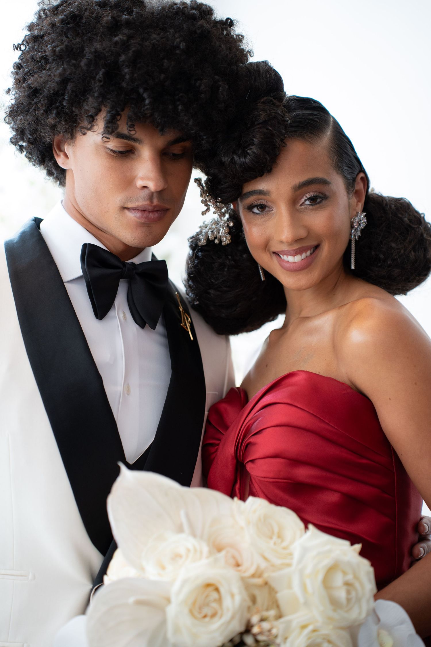 Black couple in wedding attire, bride smiling in red strapless gown holding white roses, groom in white tuxedo.