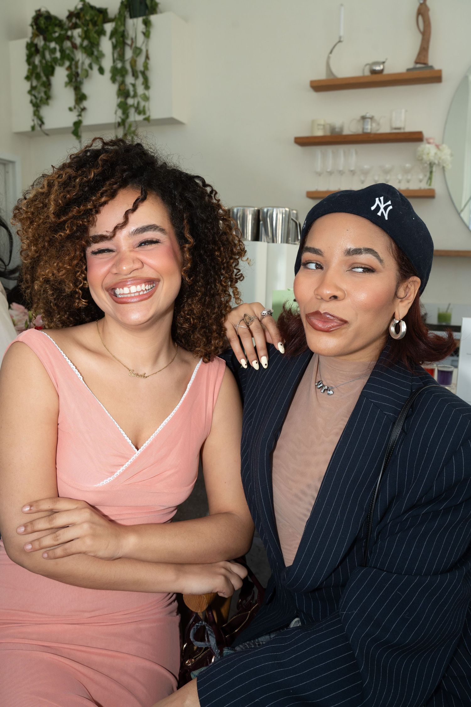Two women smile together, one in a pink dress with curly hair, one in a navy blazer.
