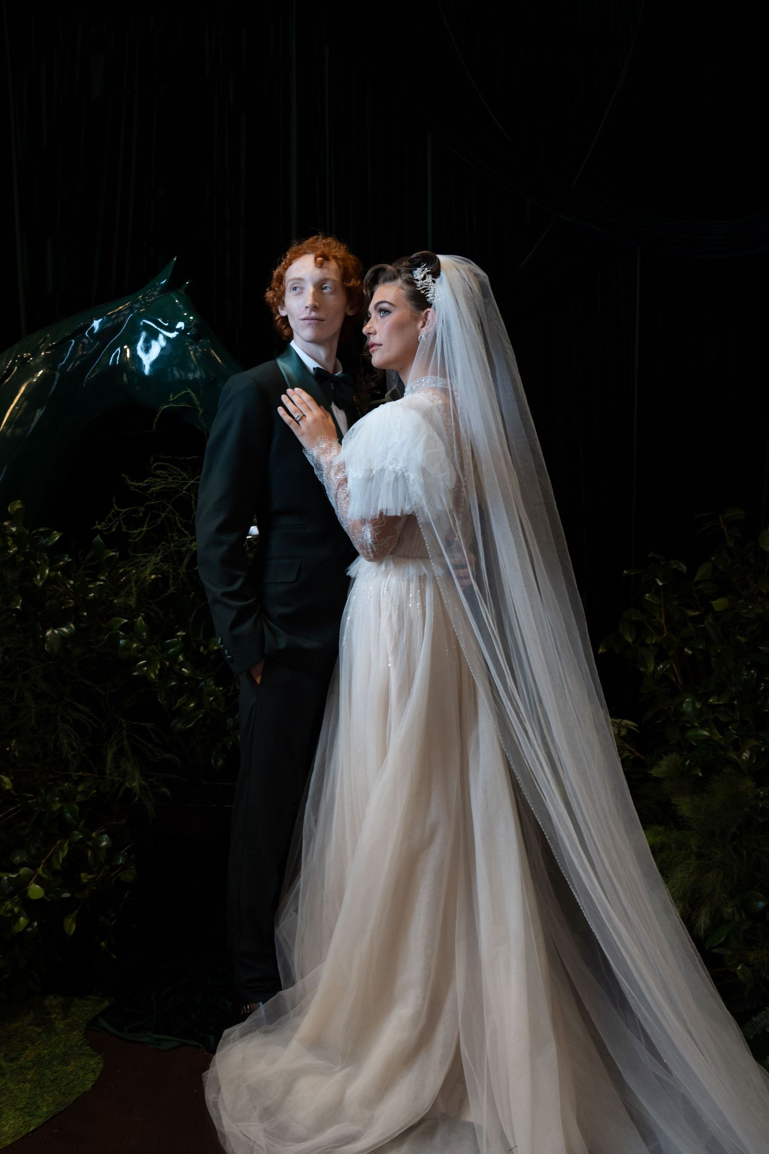 Bride in elegant tulle gown with cathedral veil stands beside red-haired groom in black suit amid dark greenery.