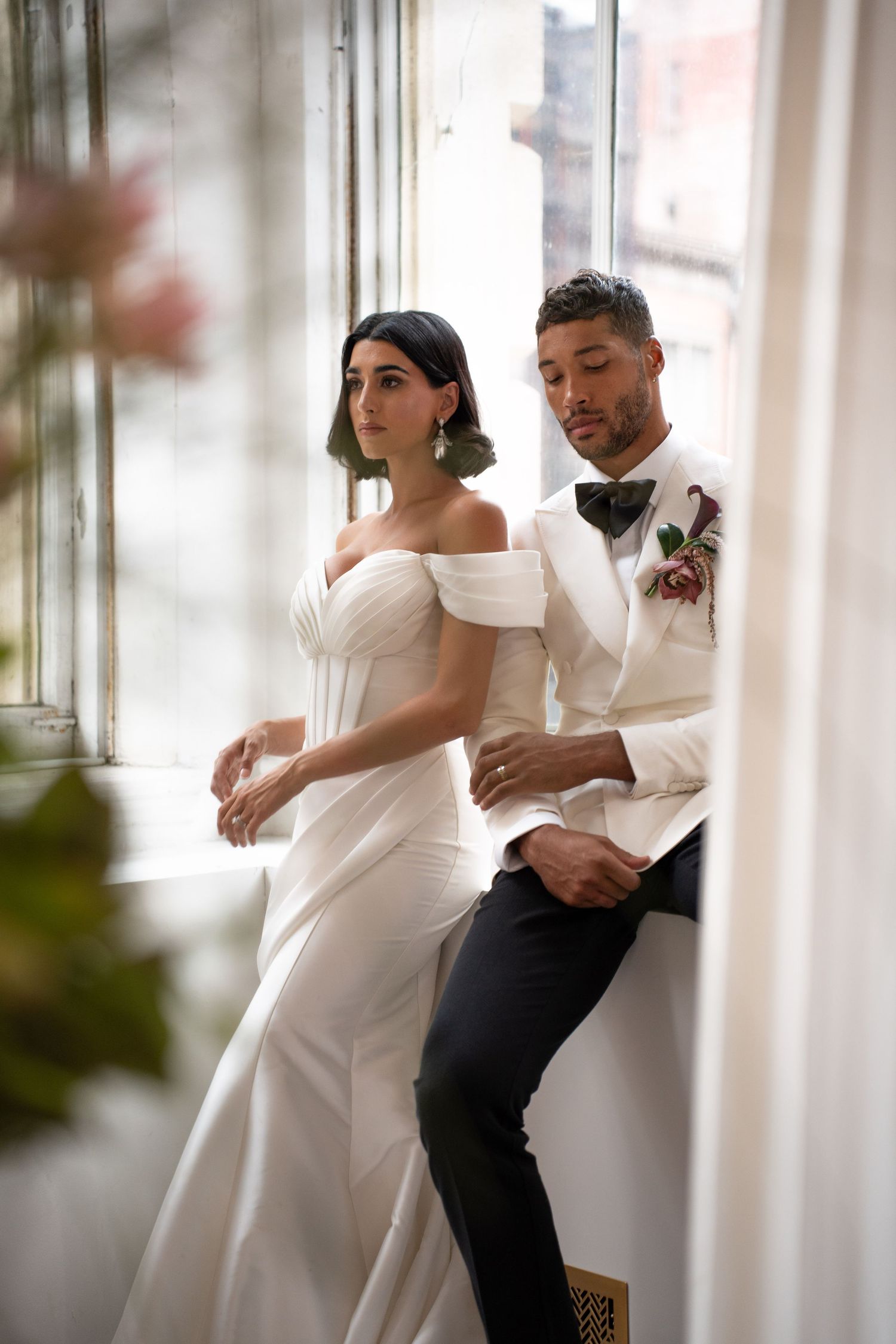 Bride in off-shoulder gown and groom in ivory tuxedo share intimate moment by sunlit window.