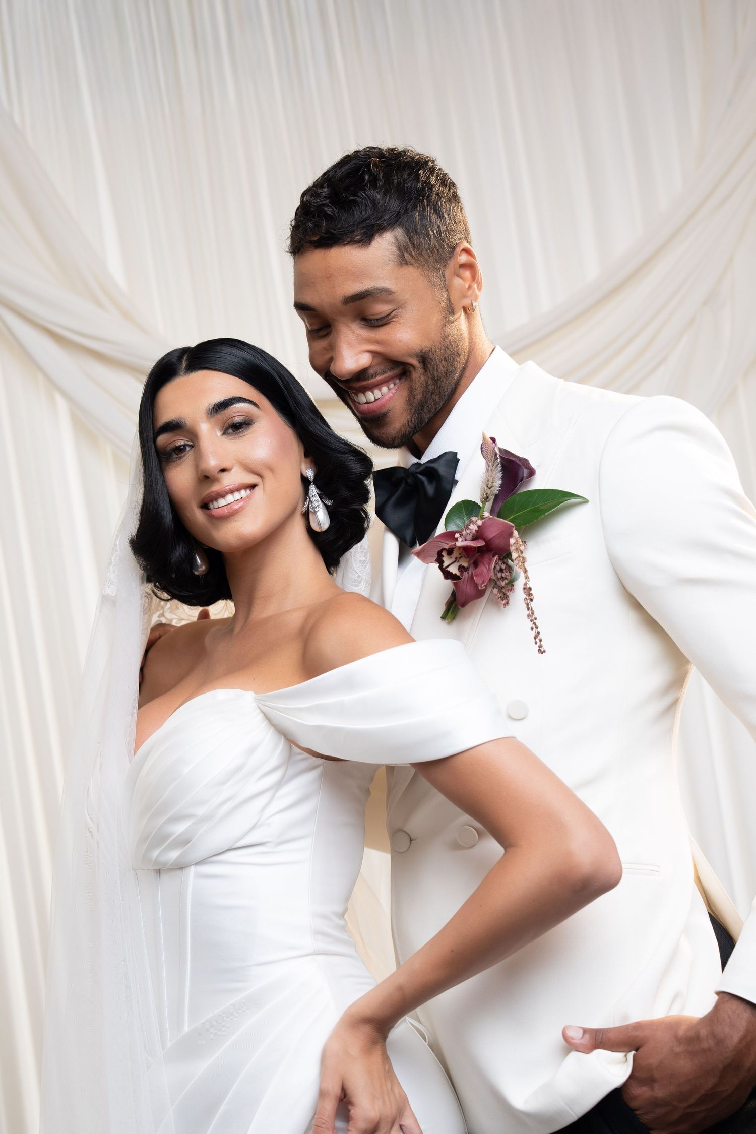 Smiling bride and groom in matching white wedding attire pose together in front of white drape backdrop.
