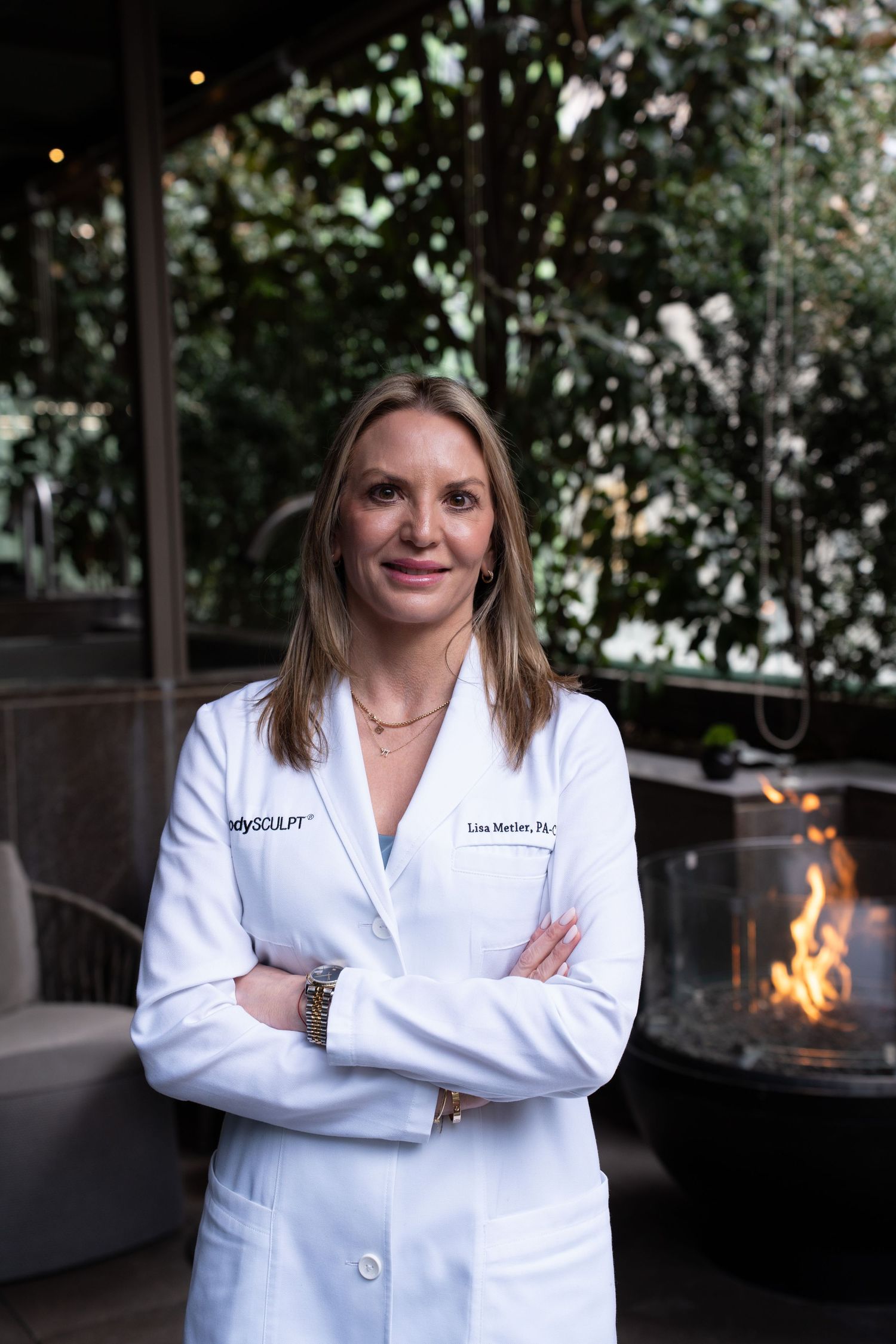 Lisa Matler PA-C in white BodySculpt coat standing confidently near an outdoor fire pit.