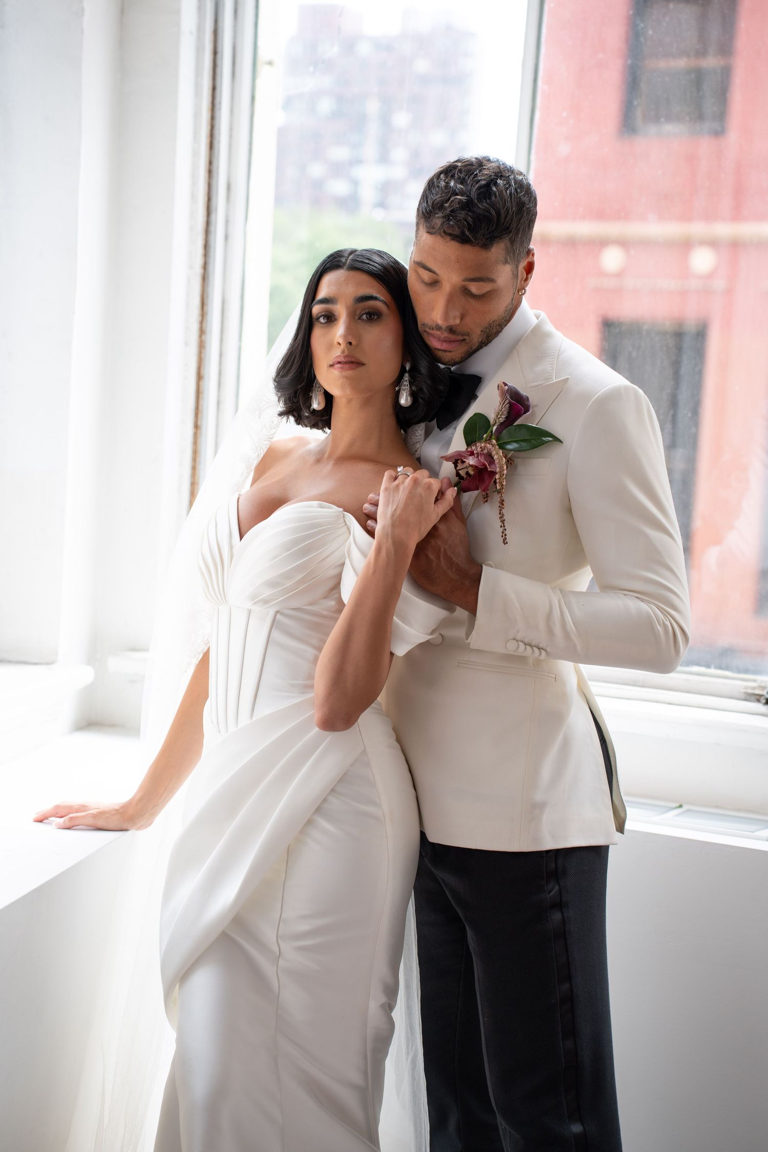 Groom in white tuxedo embracing bride in off-shoulder wedding dress by sunlit city window.