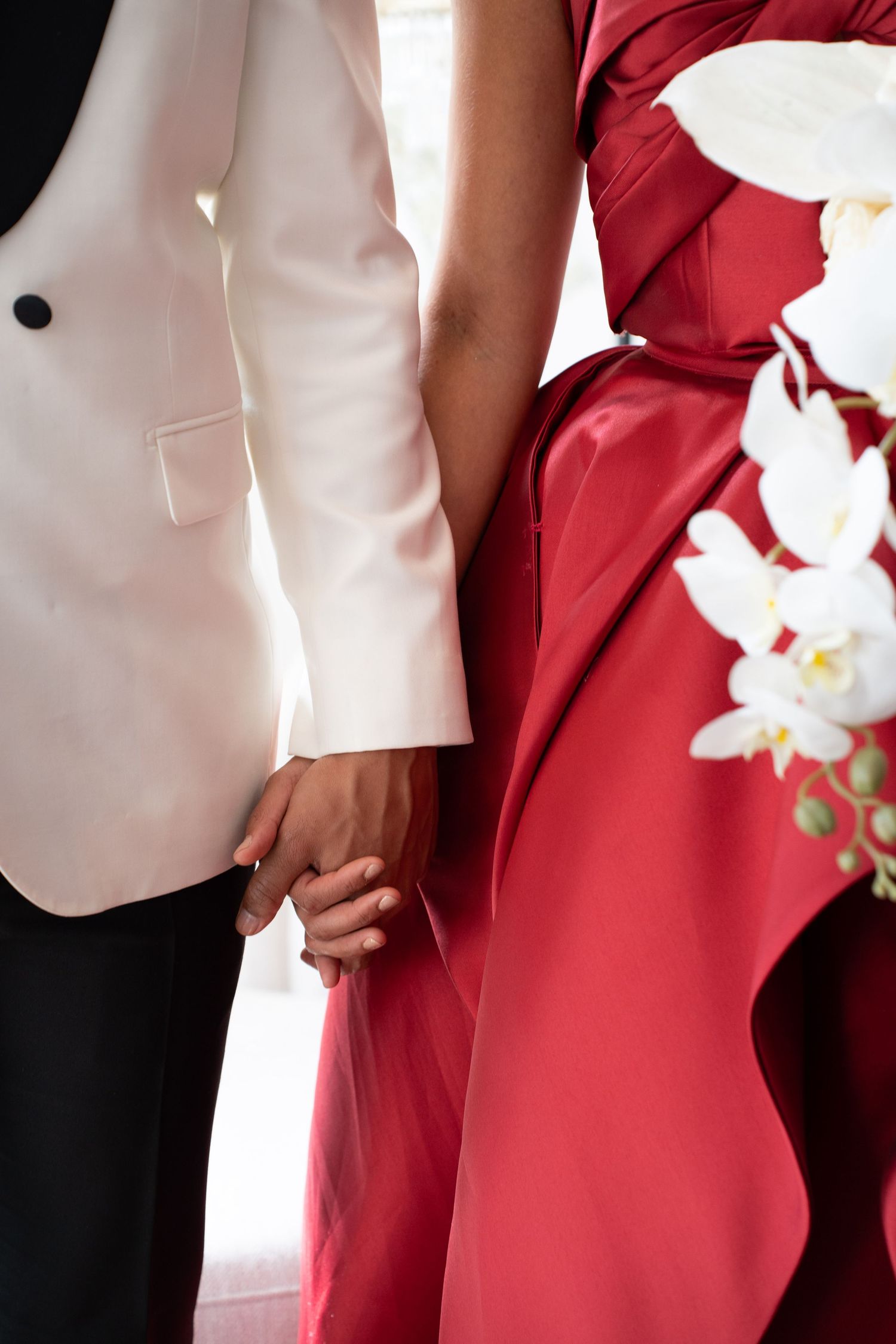 Close-up of wedding couple holding hands, groom in white tuxedo jacket, bride in red satin gown with white orchids.