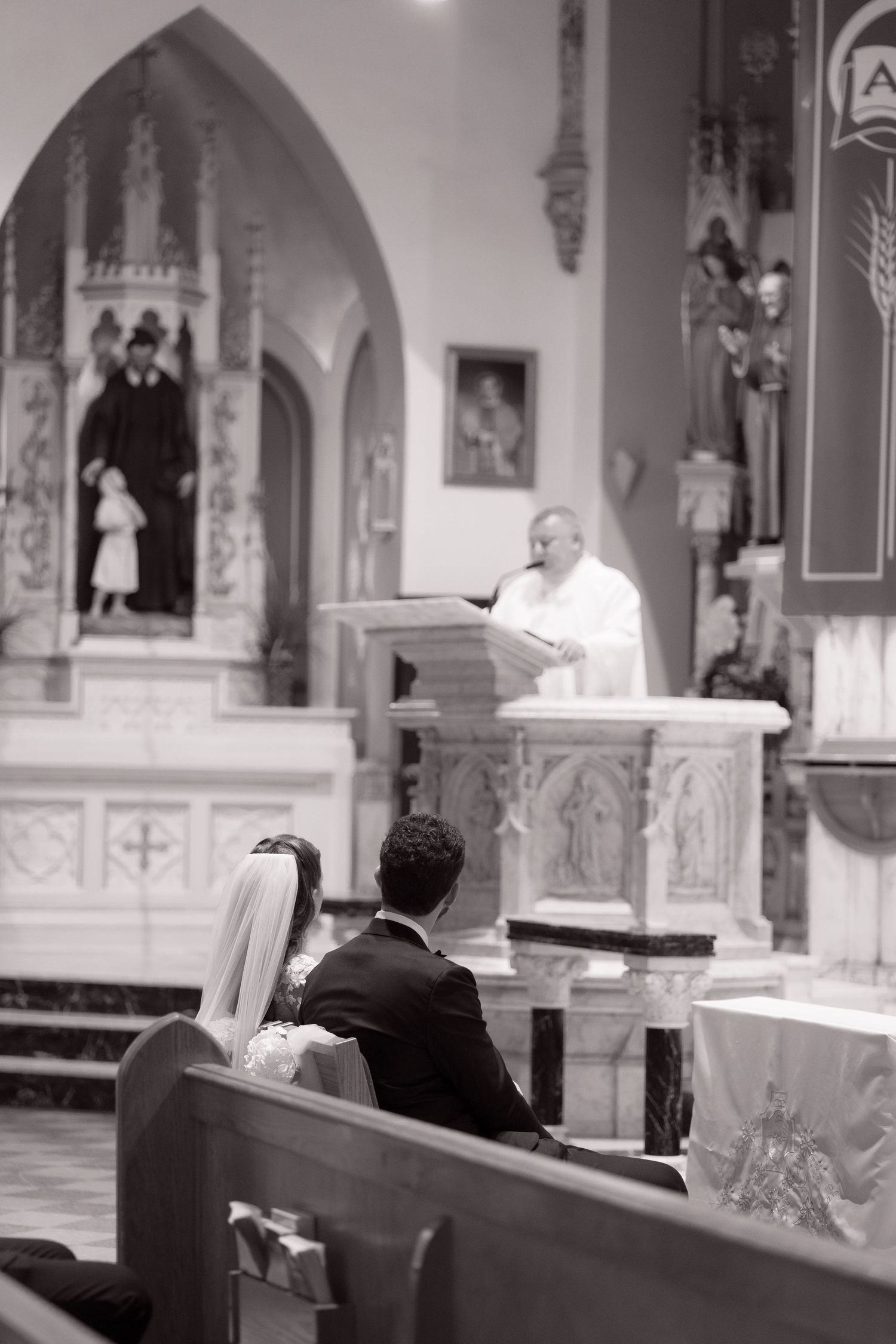 Bride and groom kneel before a priest delivering a sermon at the ornate altar during their Catholic church wedding.
