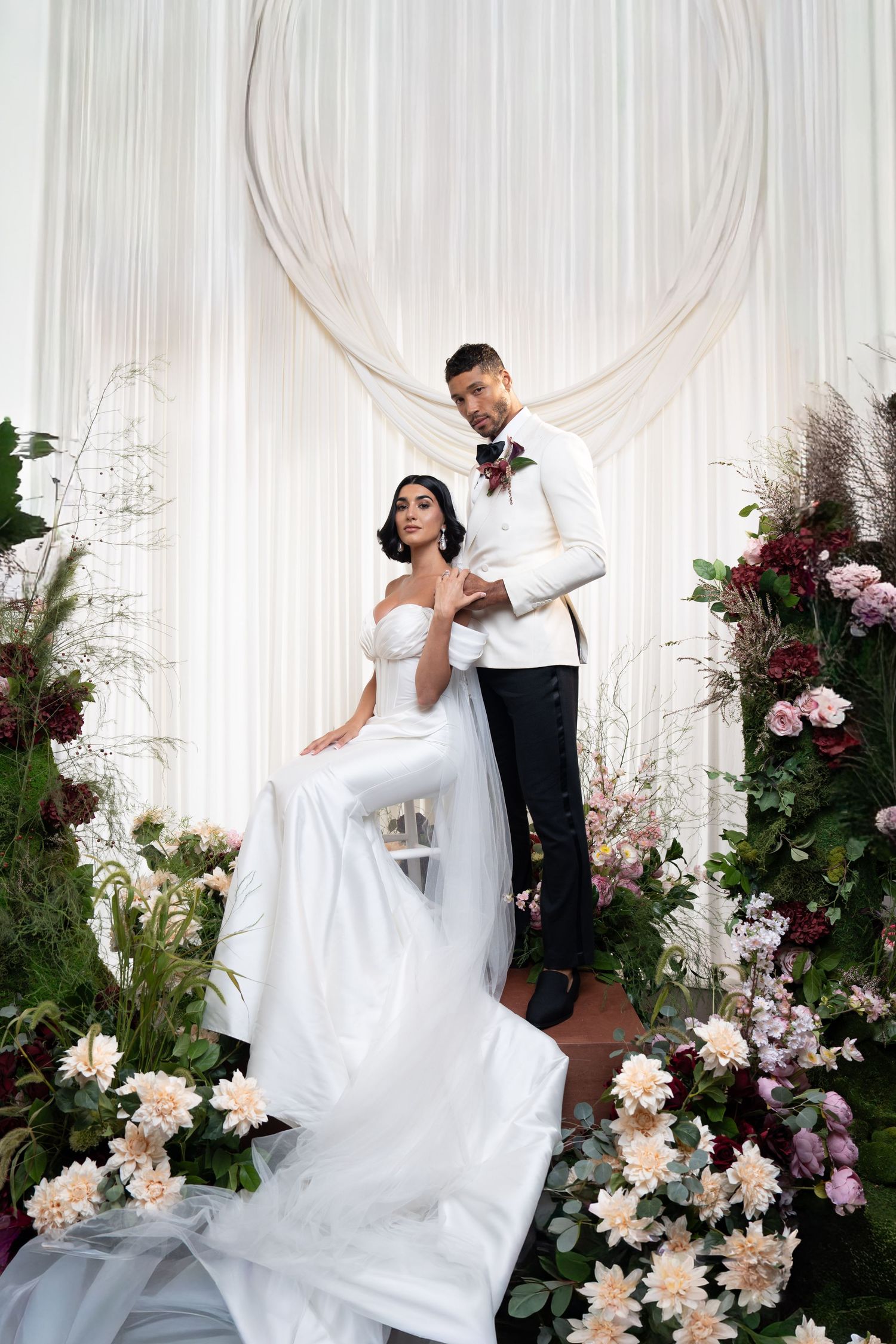 Bride in white gown and veil sits beside tall groom amid lavish floral wedding arch display.