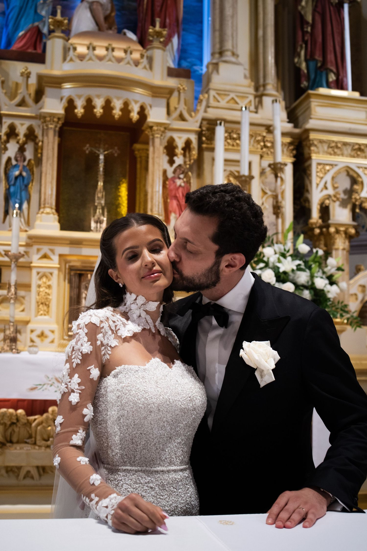 Groom kisses bride on cheek at ornate church altar, bride in lace applique wedding dress.