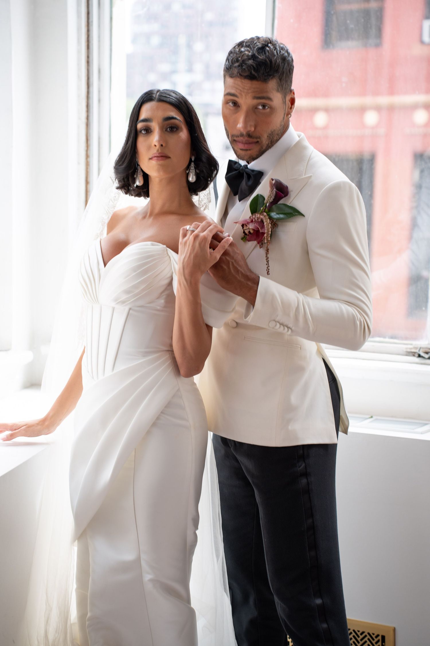 Bride in white strapless gown and groom in white tuxedo with black bow tie and floral boutonniere pose together.