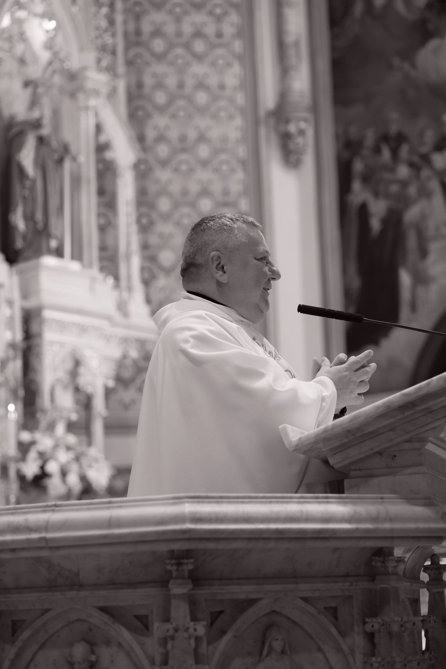 Catholic priest in white vestments delivers a wedding sermon at an ornate marble pulpit in a decorated church.