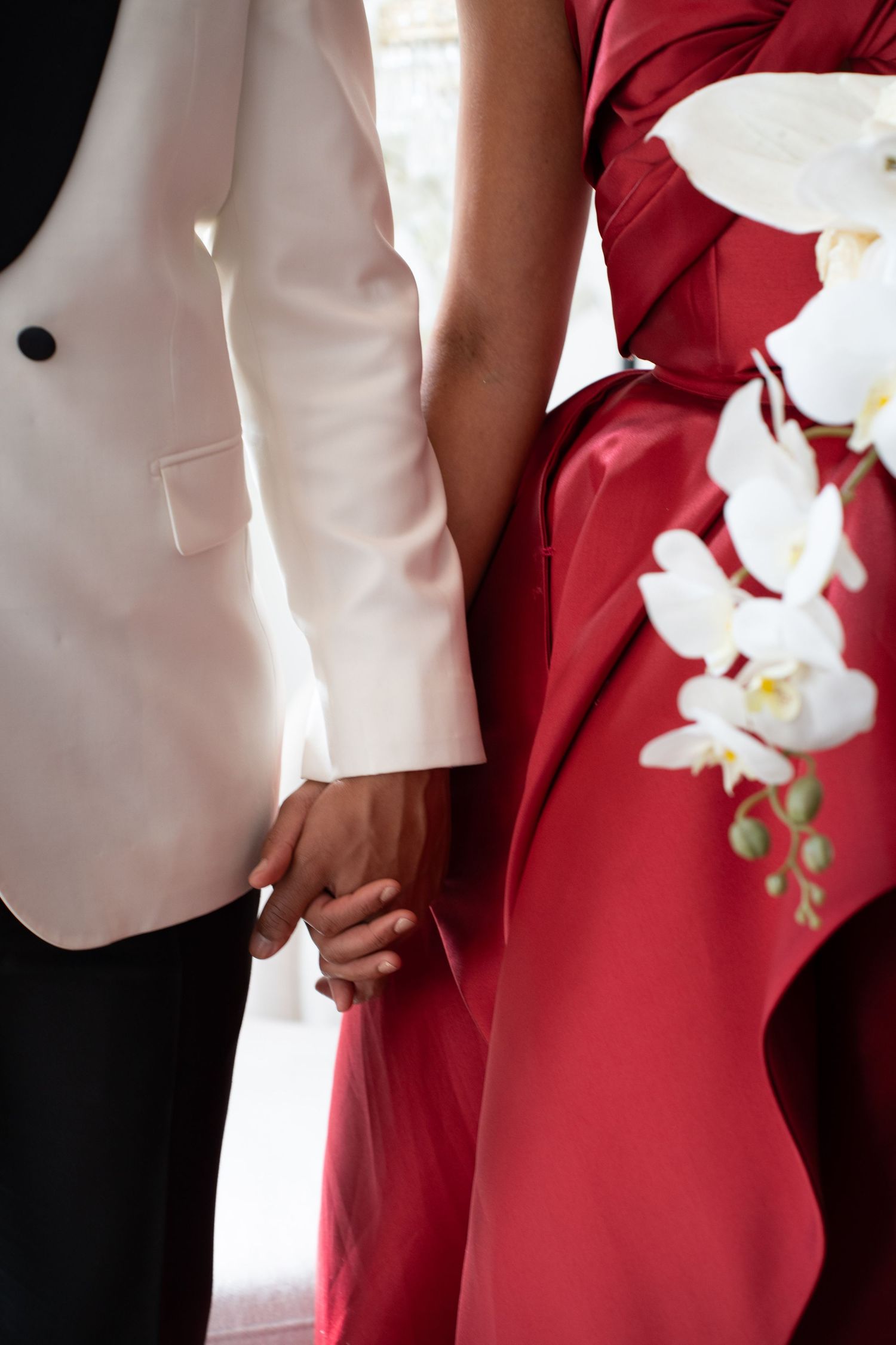 Close-up of wedding couple holding hands, groom in white tuxedo and bride in red satin gown with orchids.