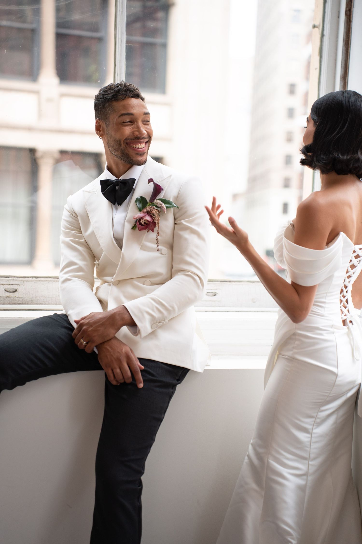 Laughing groom in white tuxedo jacket sits on windowsill chatting with bride in backless off-shoulder wedding gown.