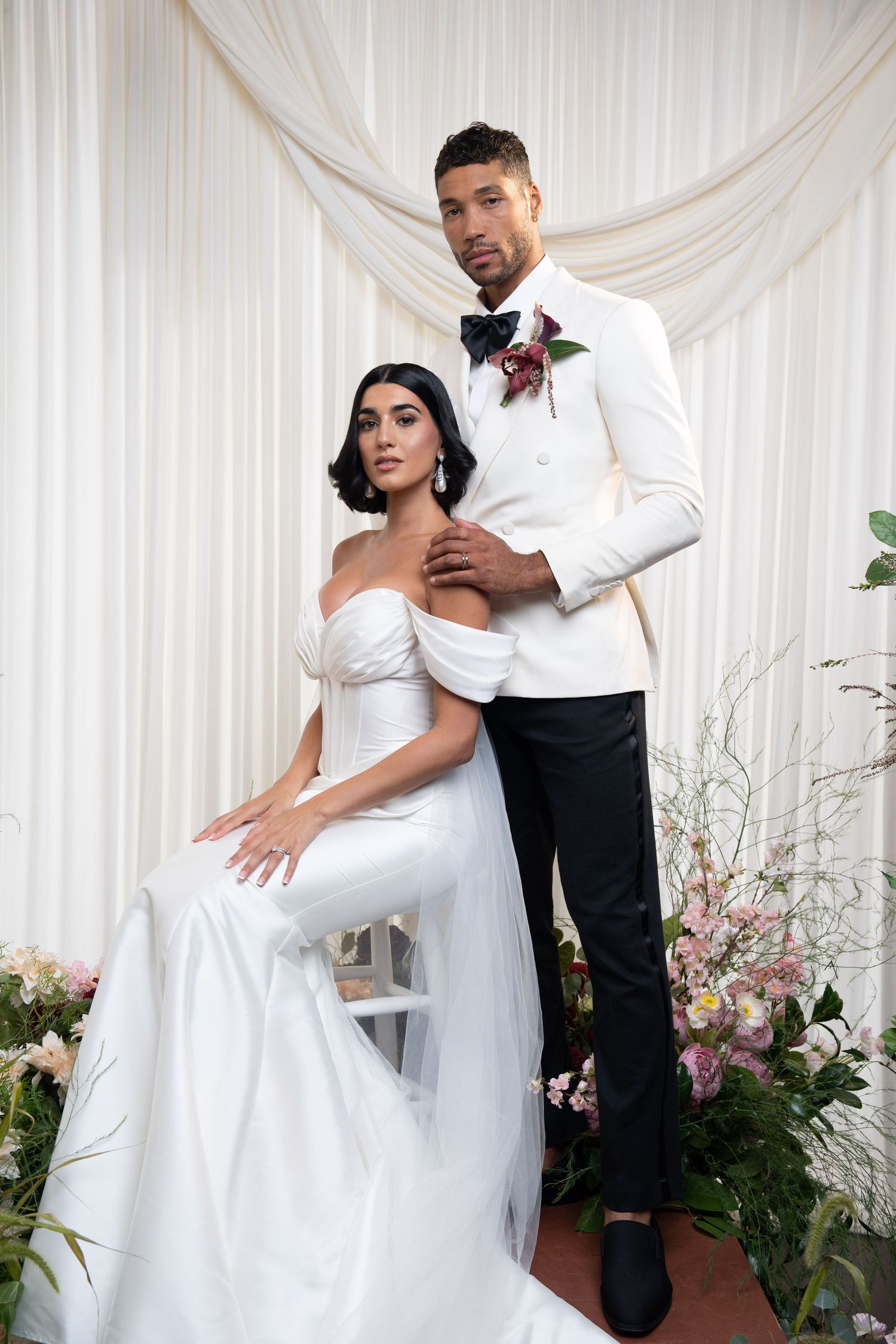 Bride in white gown sits as groom in white tuxedo stands beside her among pink florals.