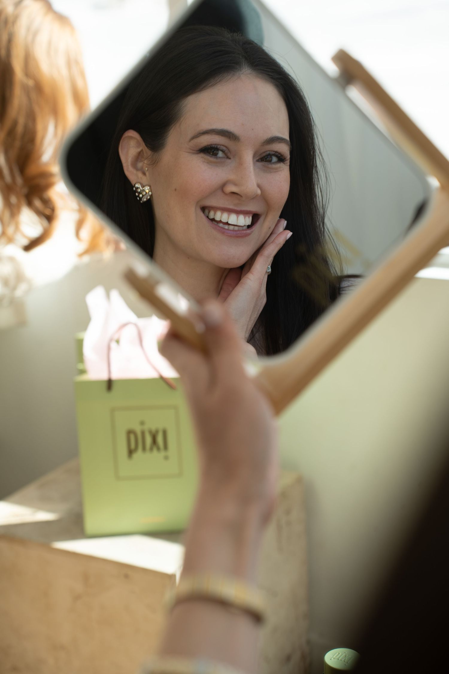 Smiling woman holding mirror reflecting her face with a green Pixi beauty bag visible behind.