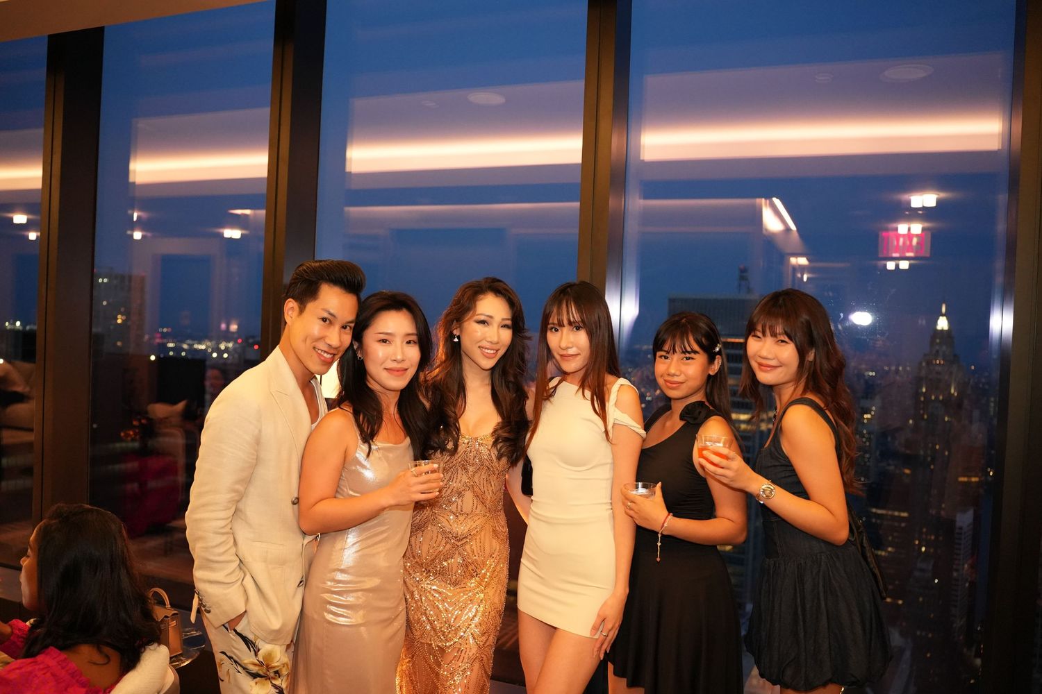 Group of six friends posing together at a rooftop party with city skyline views at dusk.